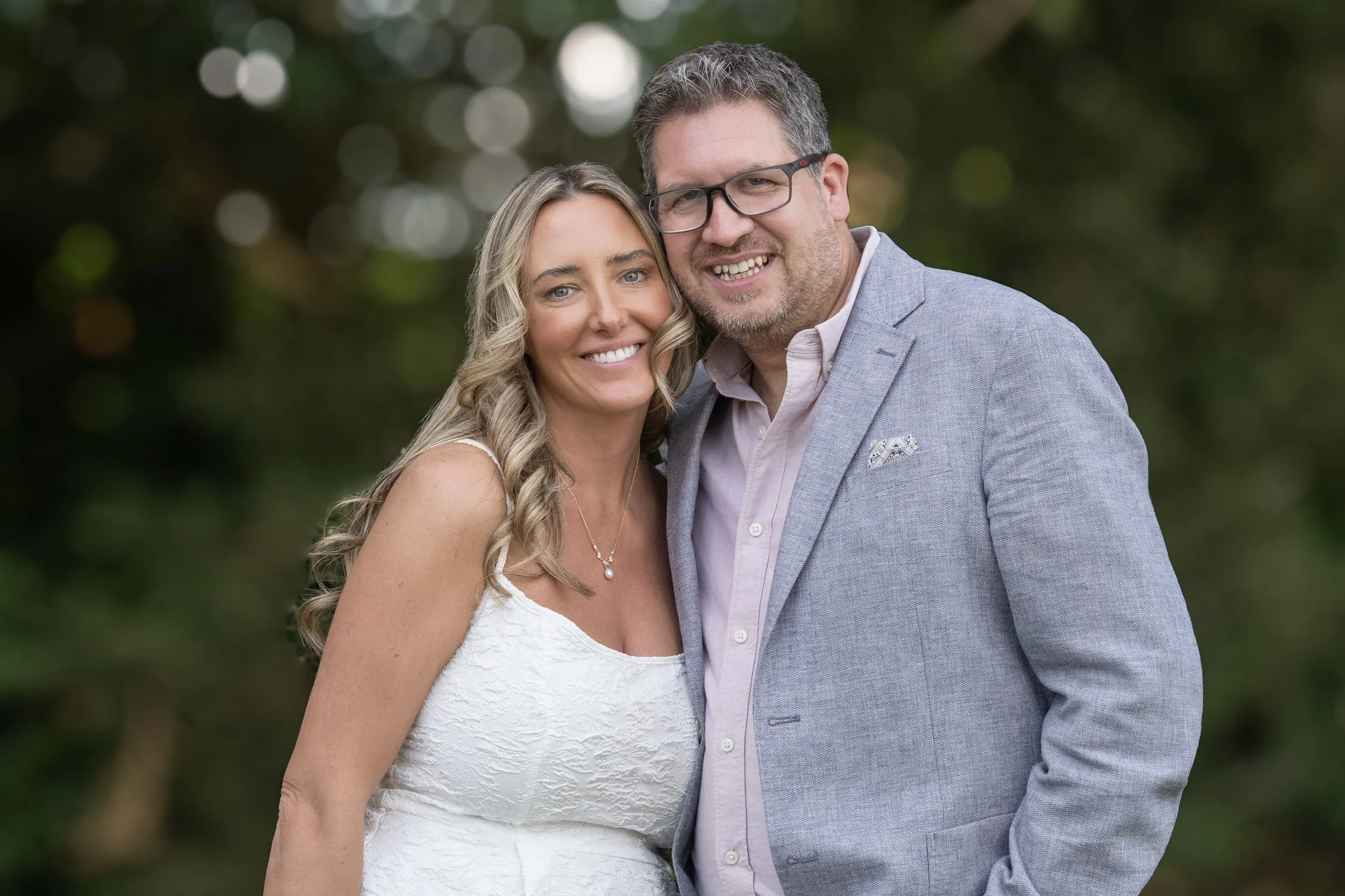 A smiling couple, a woman with long blonde hair wearing a white dress and a man with glasses and dark hair in a light gray blazer, standing close together outdoors with blurred greenery in the background.