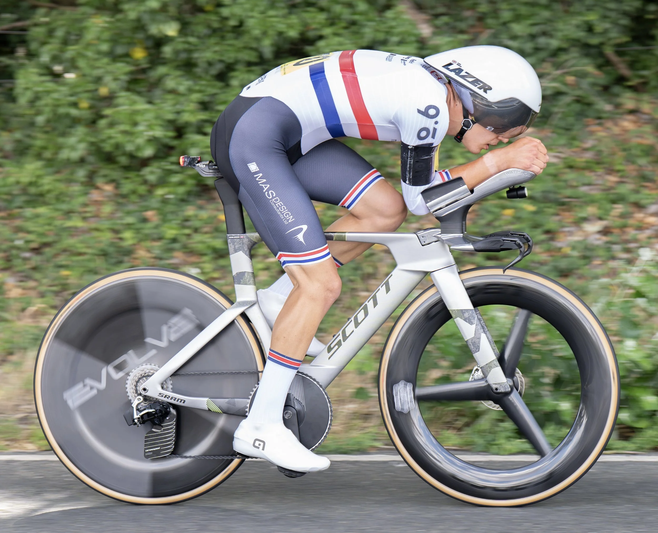 A cyclist in a white racing suit with red and blue stripes riding a road bike on pavement, with green foliage in the background.