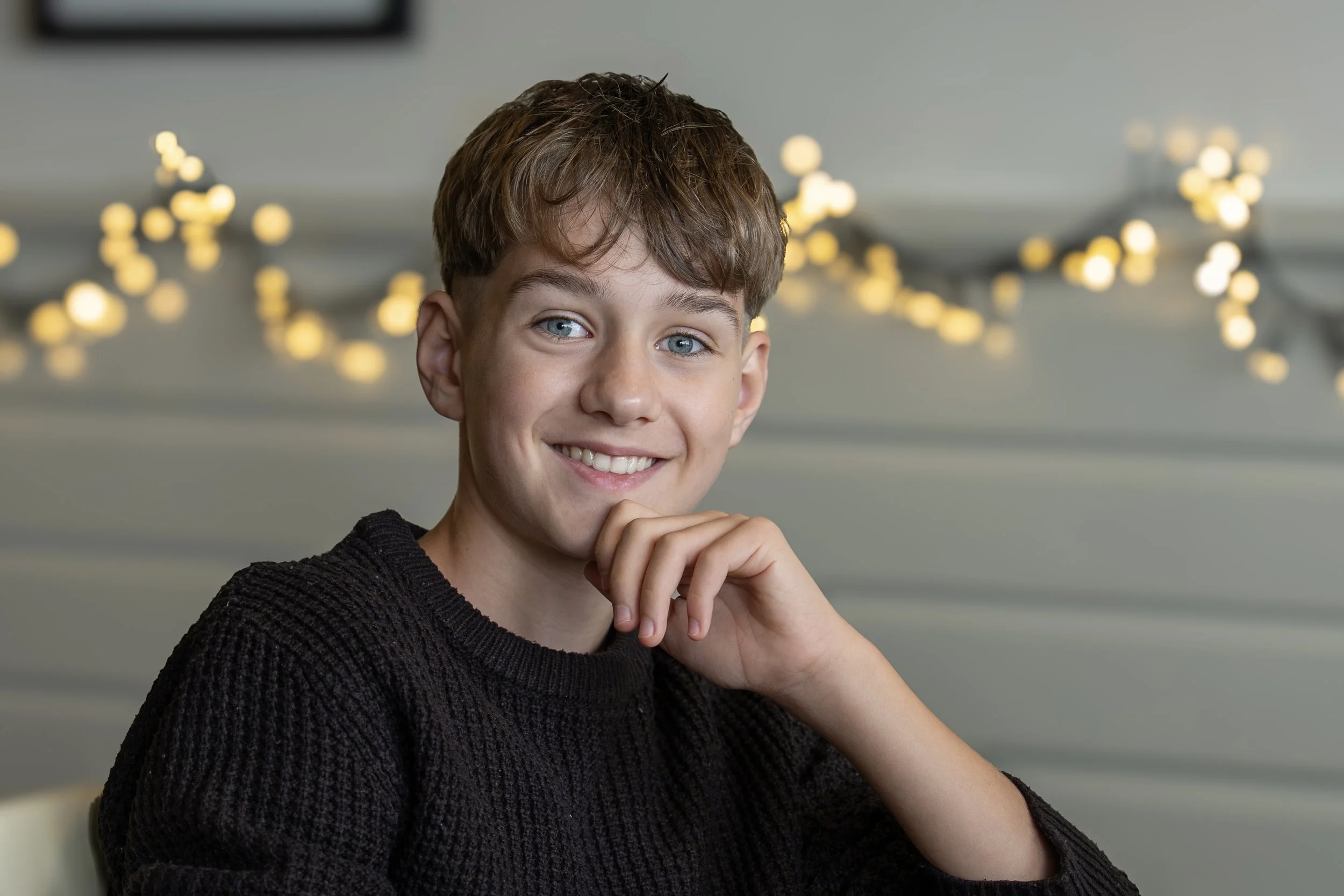 Portrait of a young man with short brown hair and blue eyes, wearing a dark sweater, smiling with his hand near his chin, standing indoors with blurry warm string lights in the background.