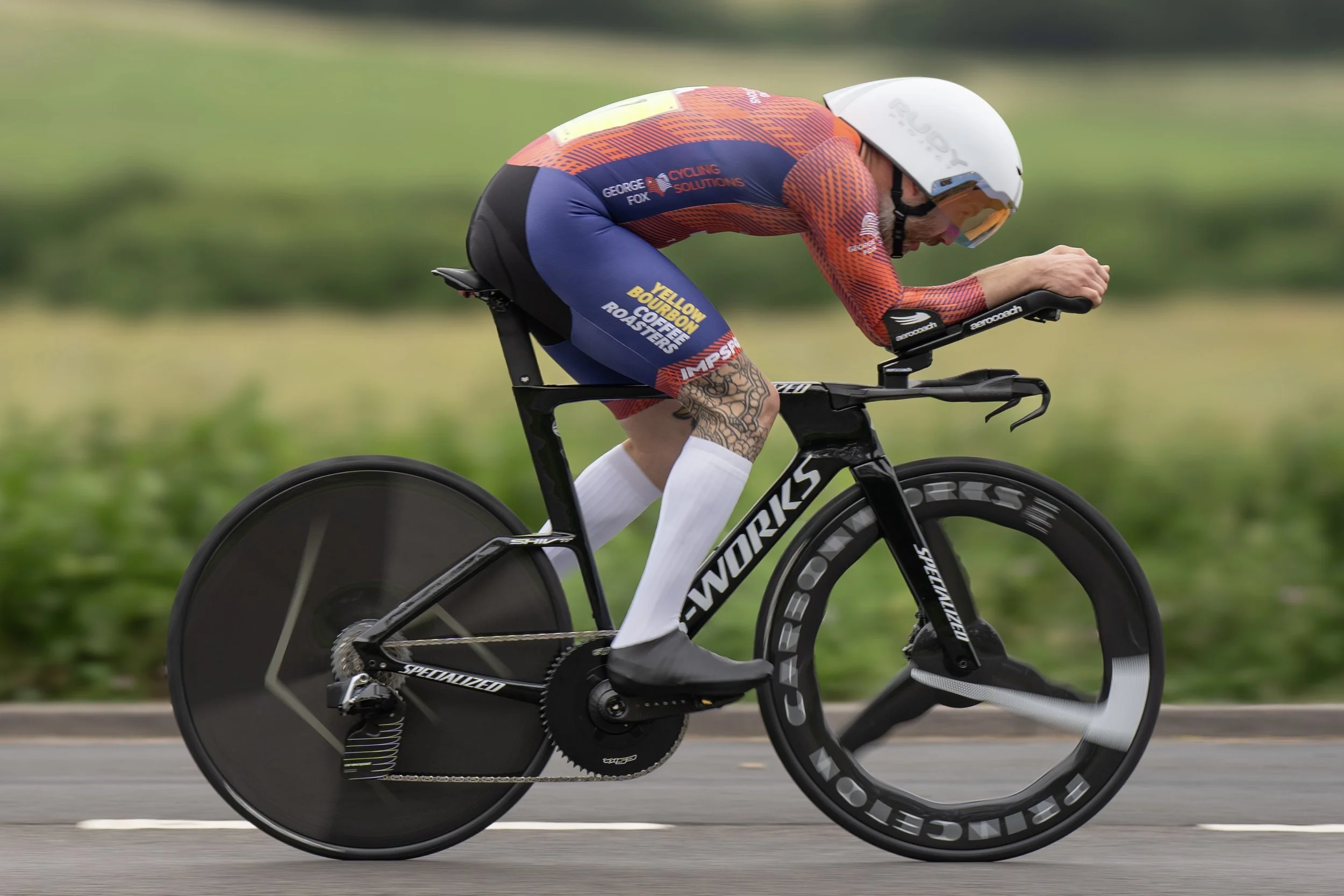 A cyclist in a racing suit with sponsors and branding, wearing a white helmet, riding a black and aerodynamic time trial bike with disc wheels on a road with blurred green background.