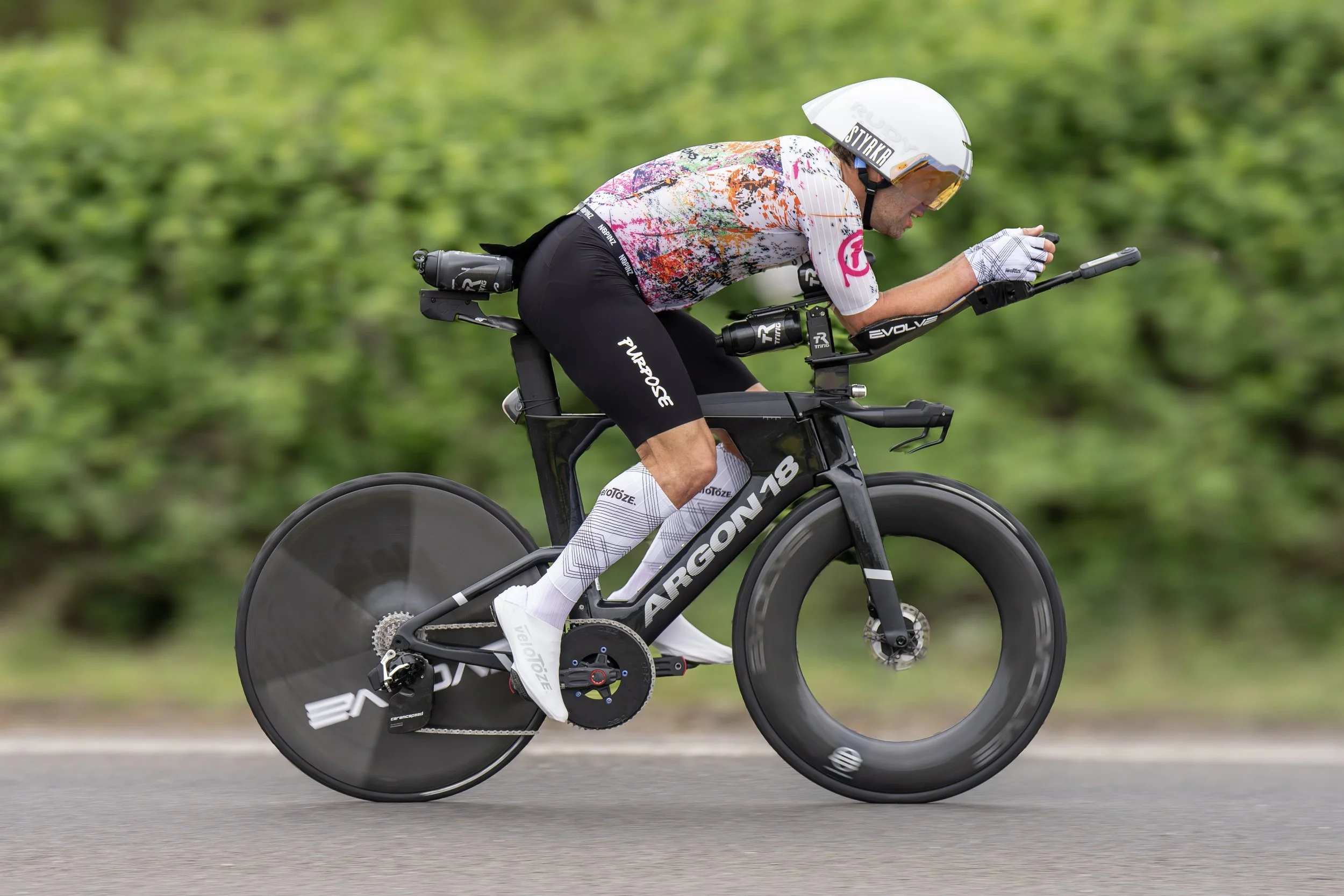 A cyclist in a colorful shirt and black shorts riding a black aerodynamic bike with disc wheels on a road with green foliage in the background.