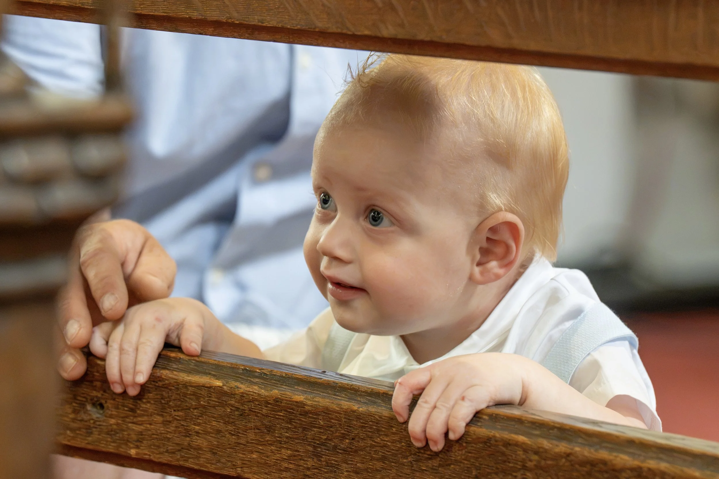 A young child with red hair looking up through a wooden railing, with hands gripping the railing in front.