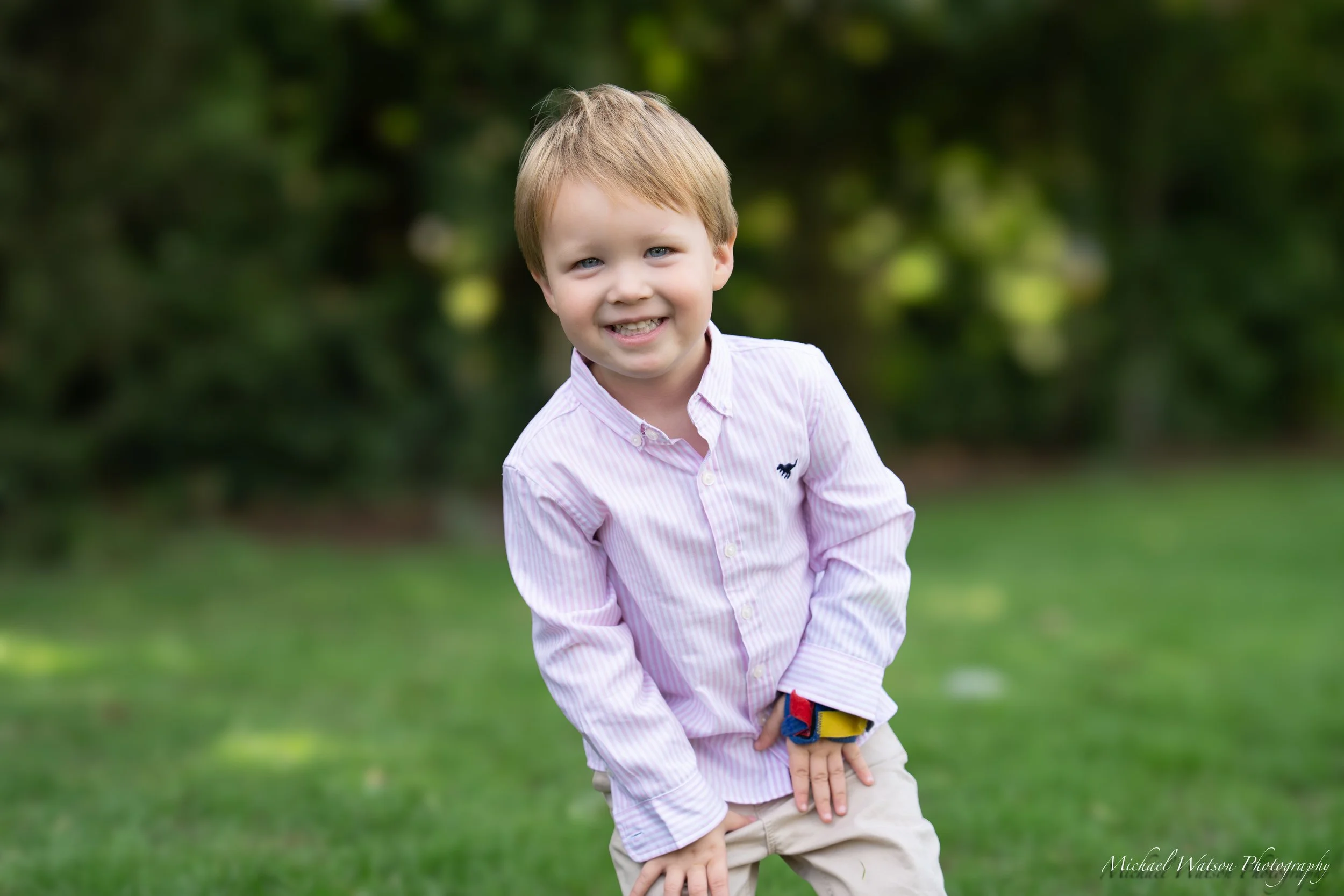 A young boy with blonde hair and blue eyes smiling outdoors, wearing a light pink striped button-up shirt with a small dark dog logo on the chest, beige pants, and a colorful wristband, standing on grass with green trees in the background.