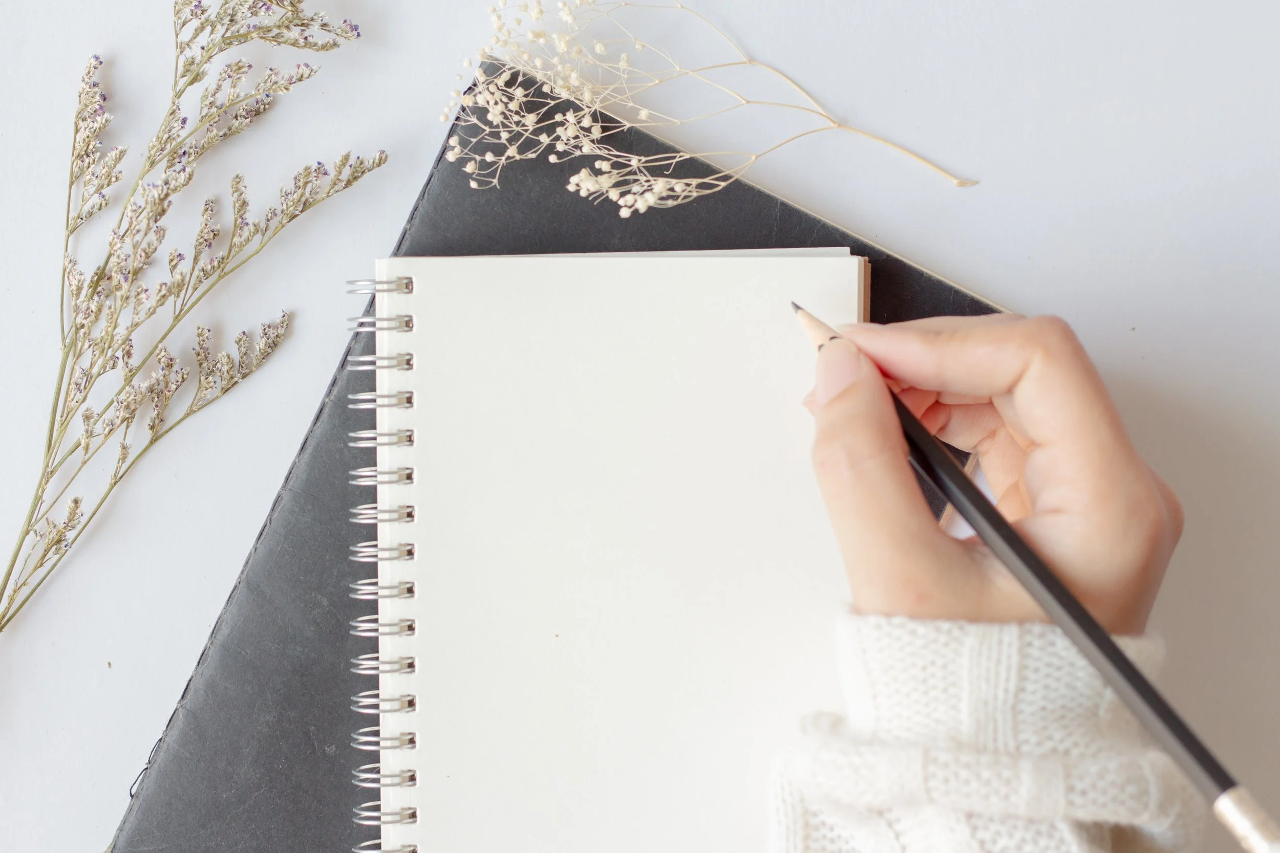A person writes in a spiral-bound notebook with a black pen, placed on top of a closed black journal or planner, surrounded by dried flowers on a white surface.