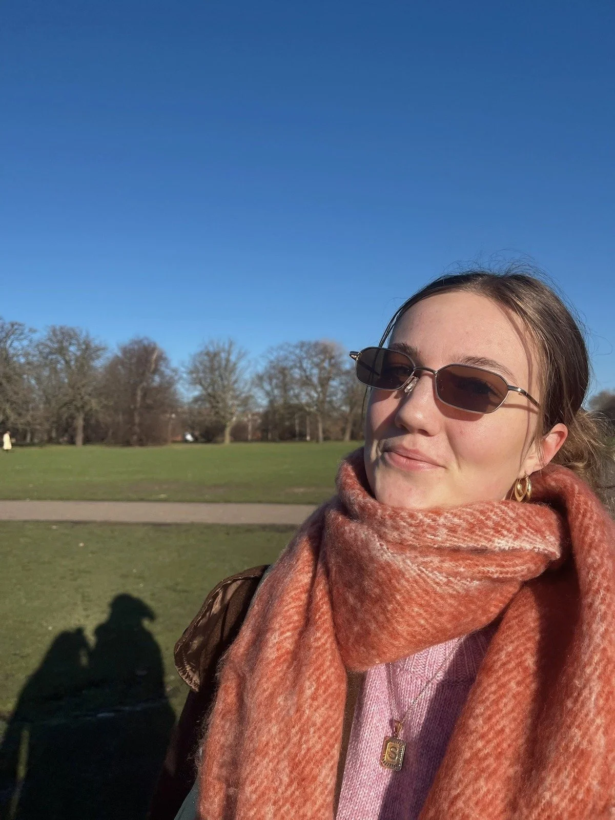 A woman wearing sunglasses, a scarf, and earrings, smiling outdoors on a sunny day with trees and grass in the background.