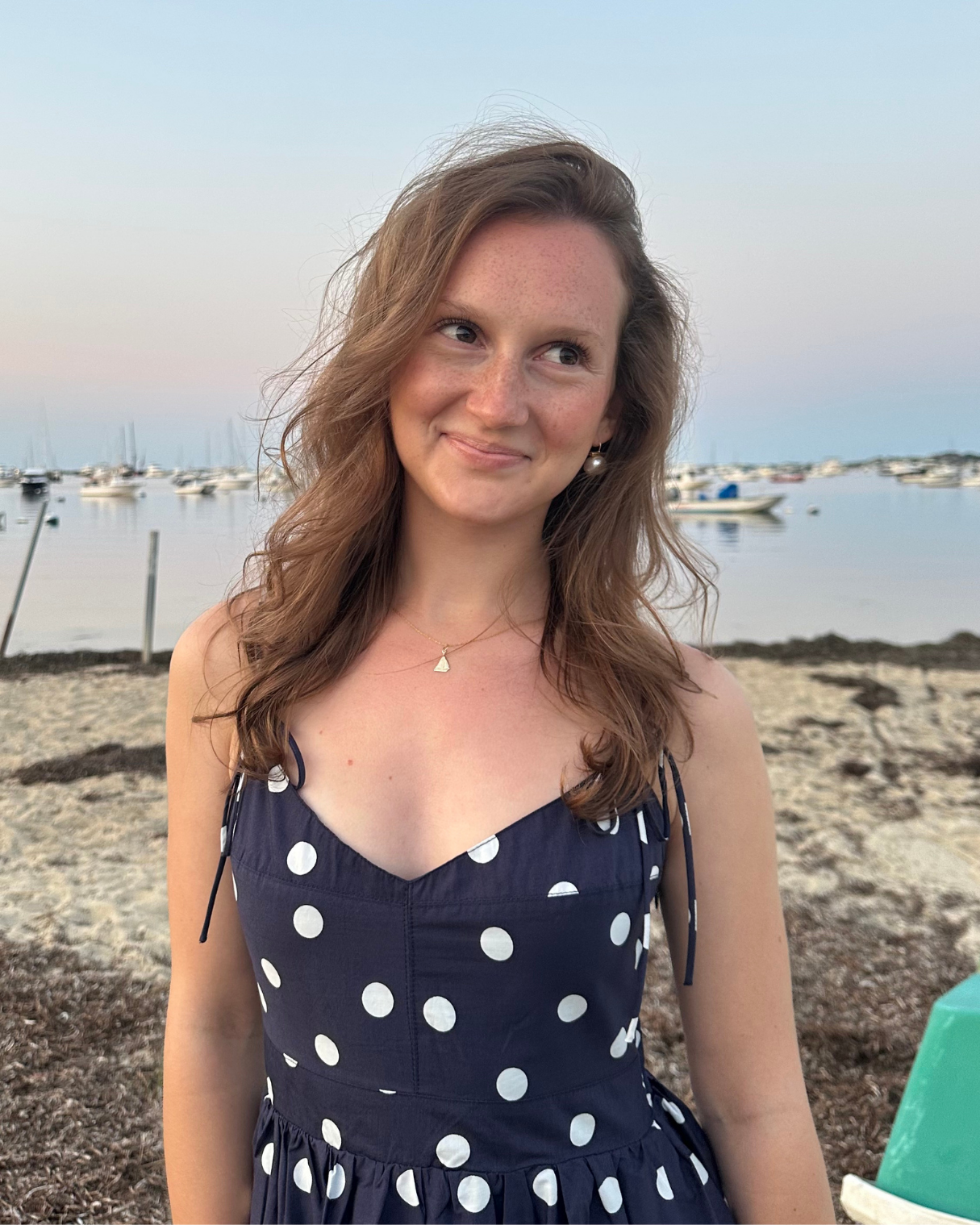 A young woman with shoulder-length brown hair, in a navy blue dress with white polka dots, standing on a beach at sunset, with boats anchored on calm water in the background.