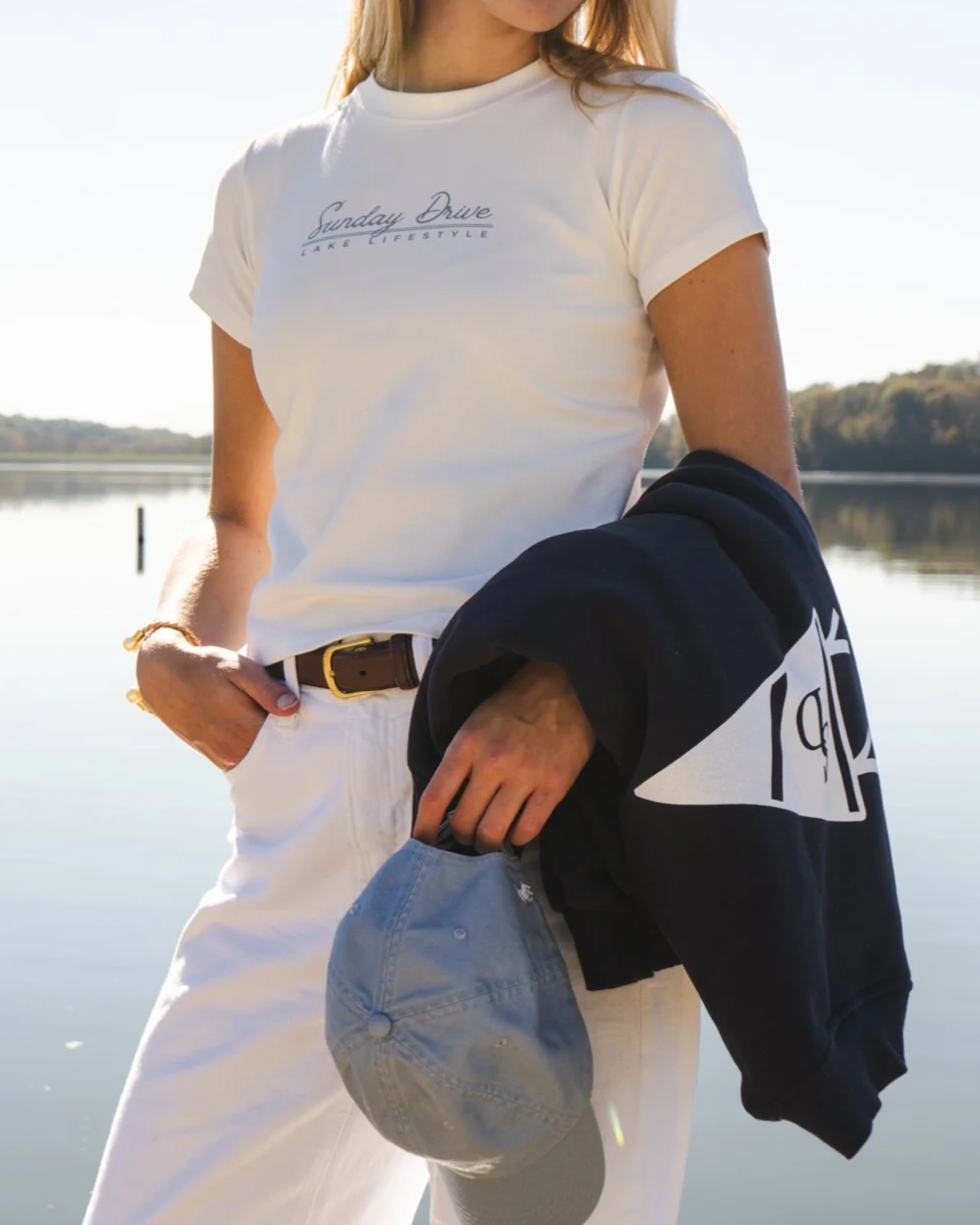 A woman standing near a lake holding a navy blue hoodie and a gray baseball cap, wearing a white t-shirt with text, with trees and water in the background.