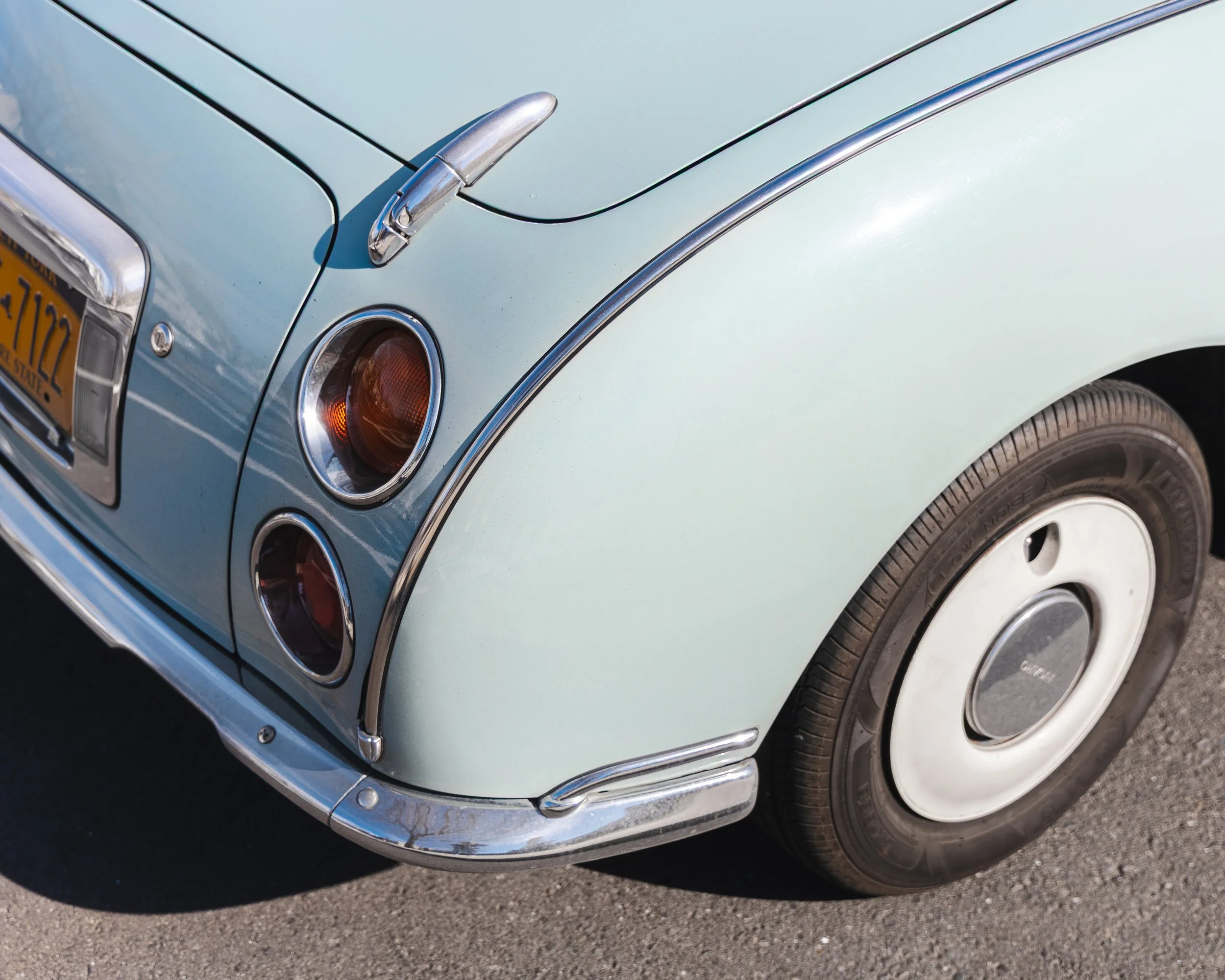 Close-up of the front corner of a vintage light blue car, showing the headlight, turn signal, chrome bumper, and part of the hood with a chrome detail and a vintage license plate.