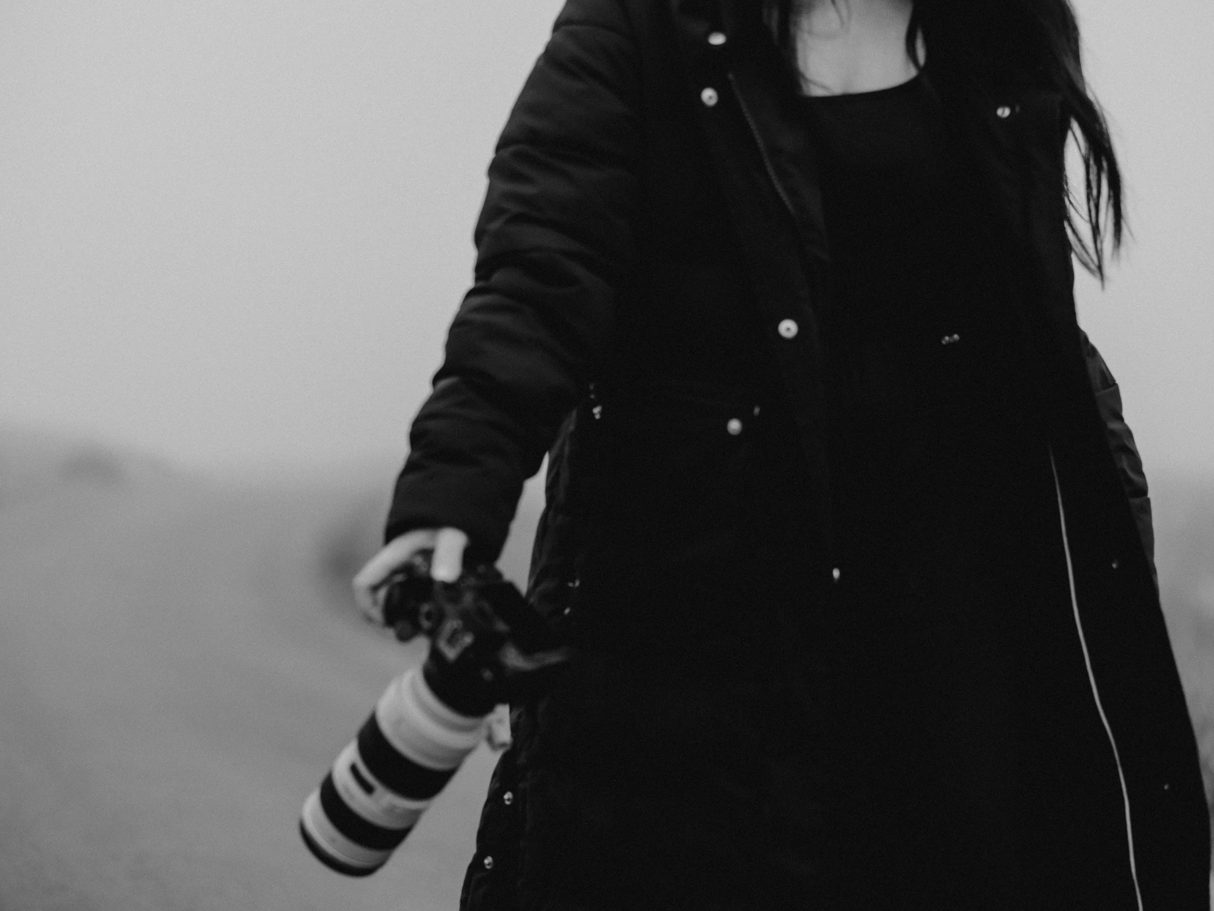 A person wearing a black jacket and striped arm warmers, holding a tennis racket on a cloudy day.