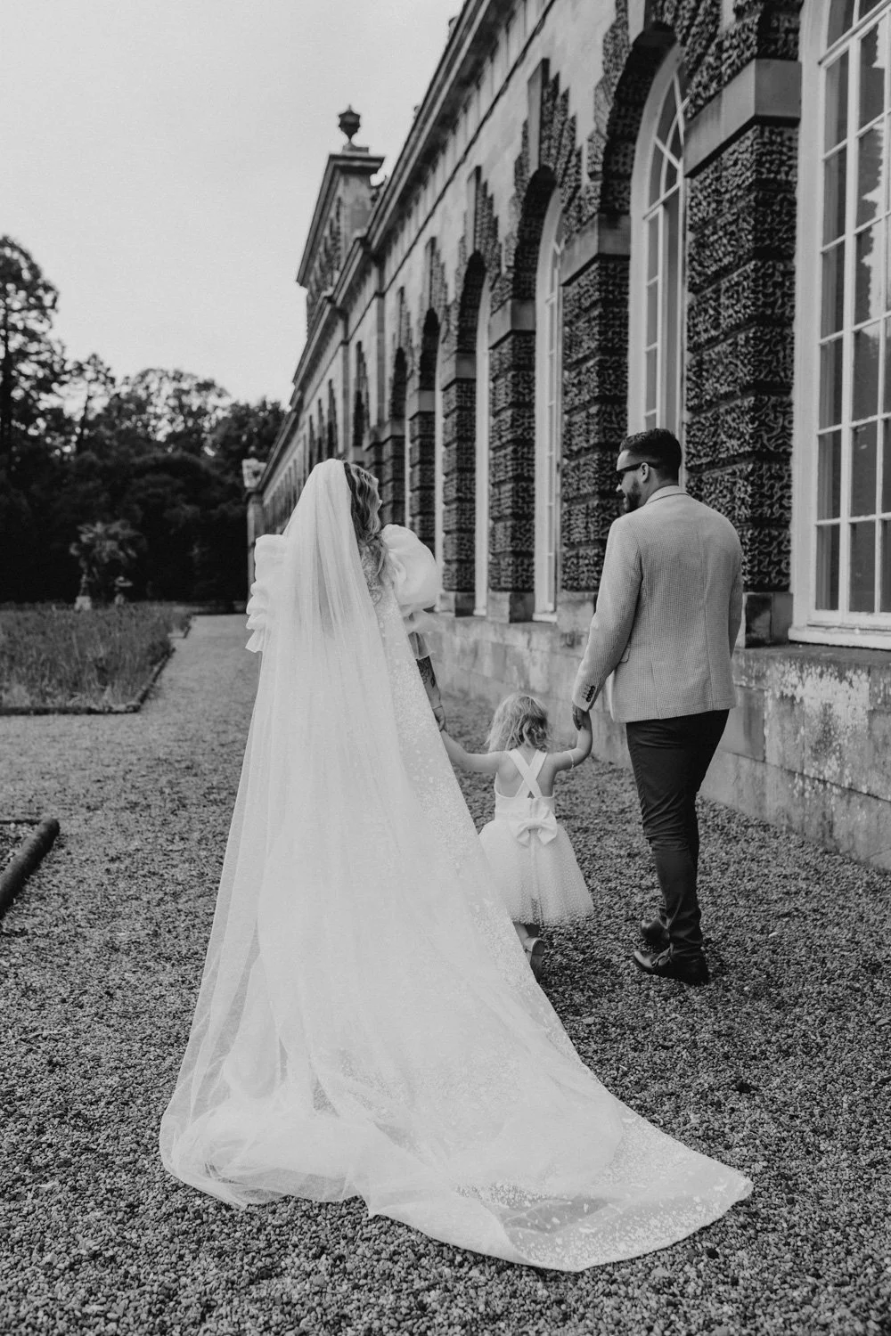 A wedding scene in black and white with a bride in a long veil and gown, a groom, and two young girls walking hand in hand outside a historic building with large windows.