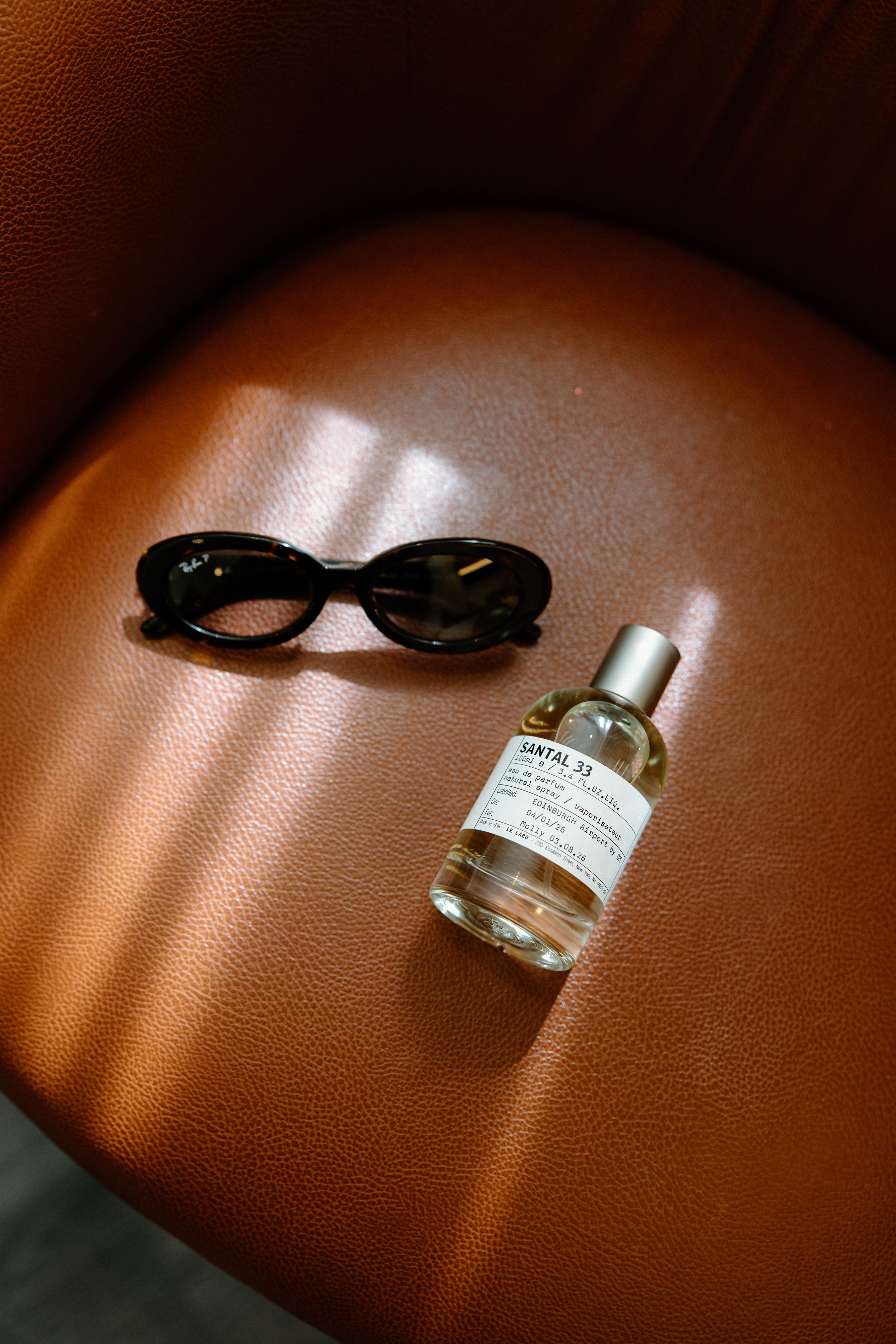 A pair of black sunglasses and a small bottle of perfume placed on a textured, reddish-brown leather surface.