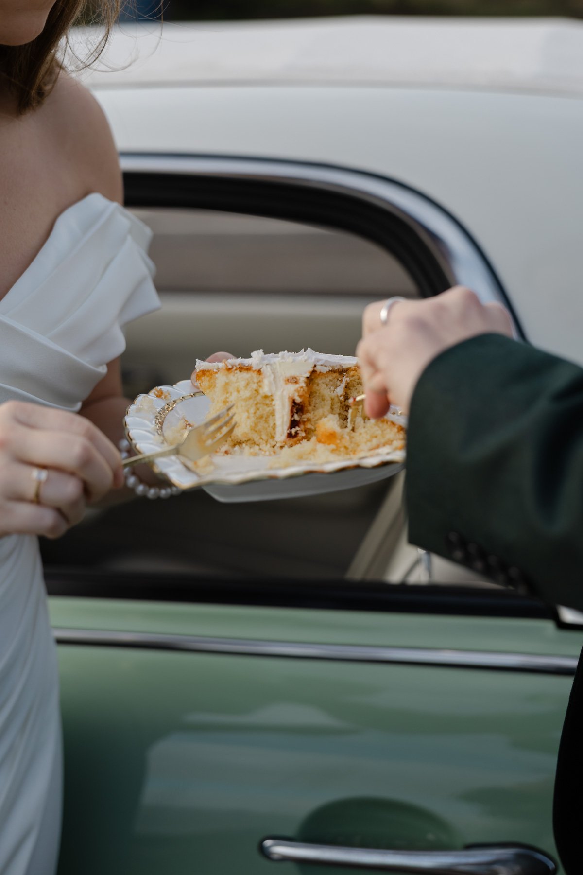 A person wearing a white dress and face mask is holding a plate with a slice of cake, handing it through the window of a car to someone wearing a dark suit.