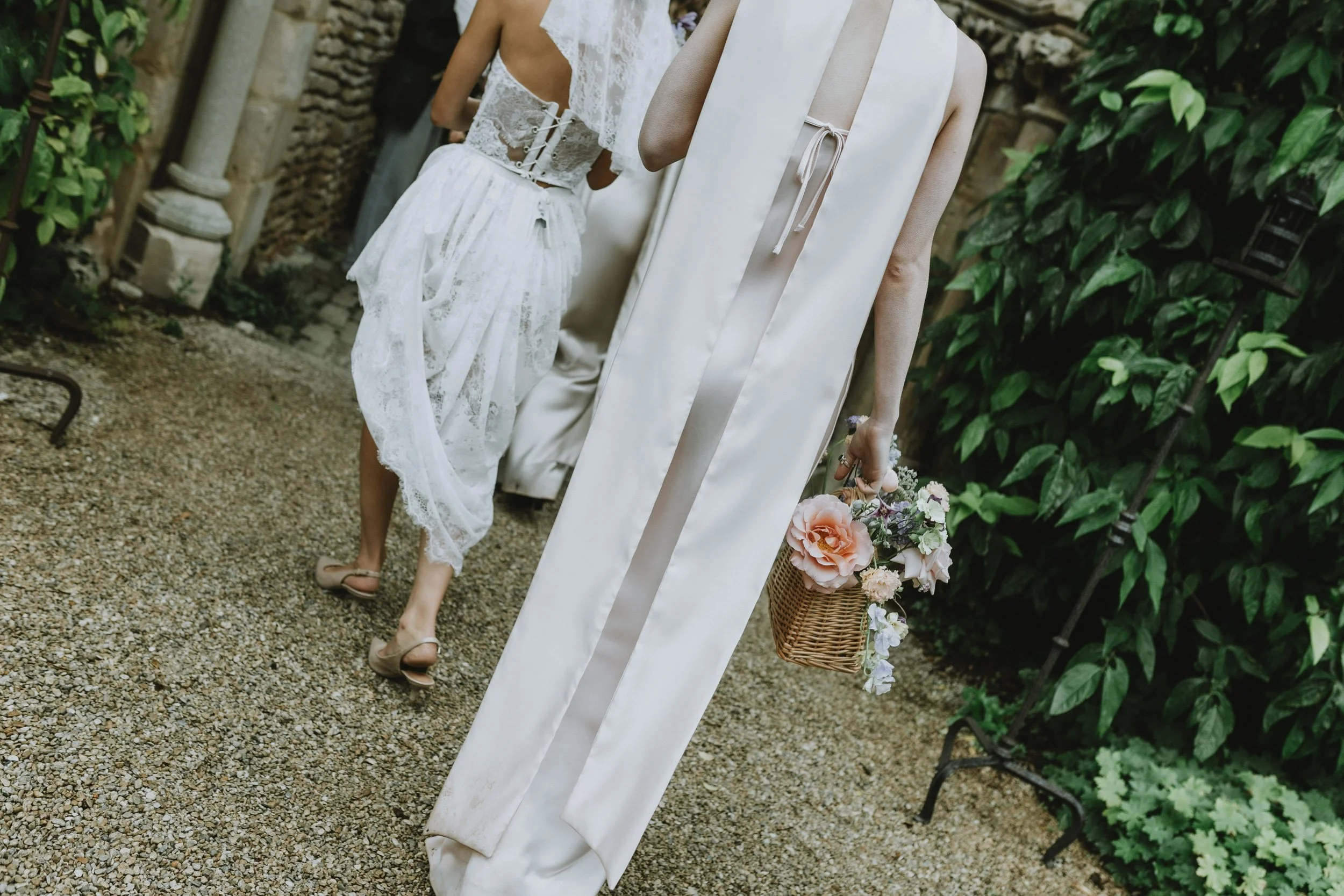 Women dressed in white wedding attire walking outdoors on a gravel path with greenery and stone wall in the background. One woman is carrying a basket of flowers.