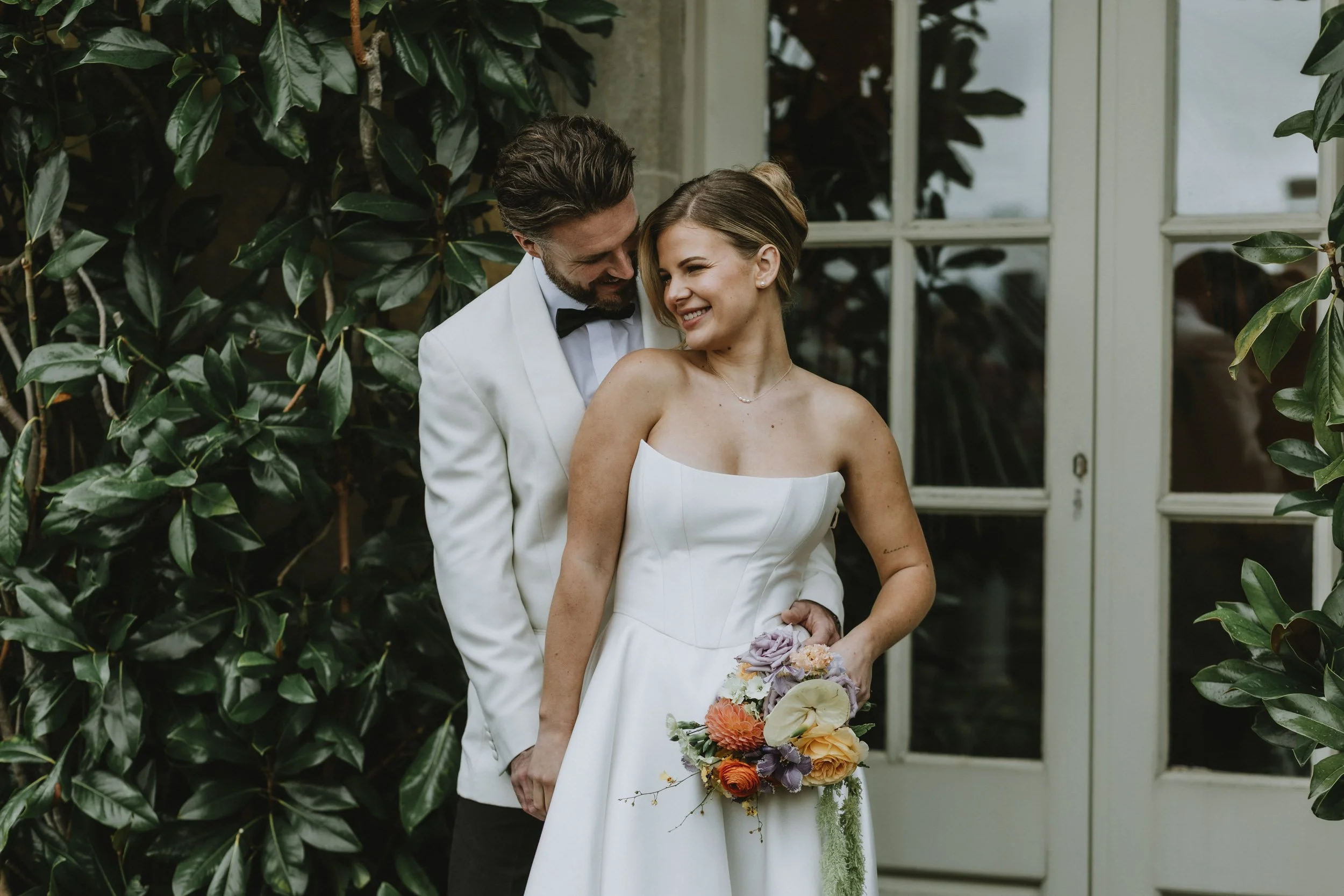 A newlywed couple in wedding attire standing close together outdoors in front of a green leafy backdrop and glass door, with the groom leaning his head toward the bride and both smiling.