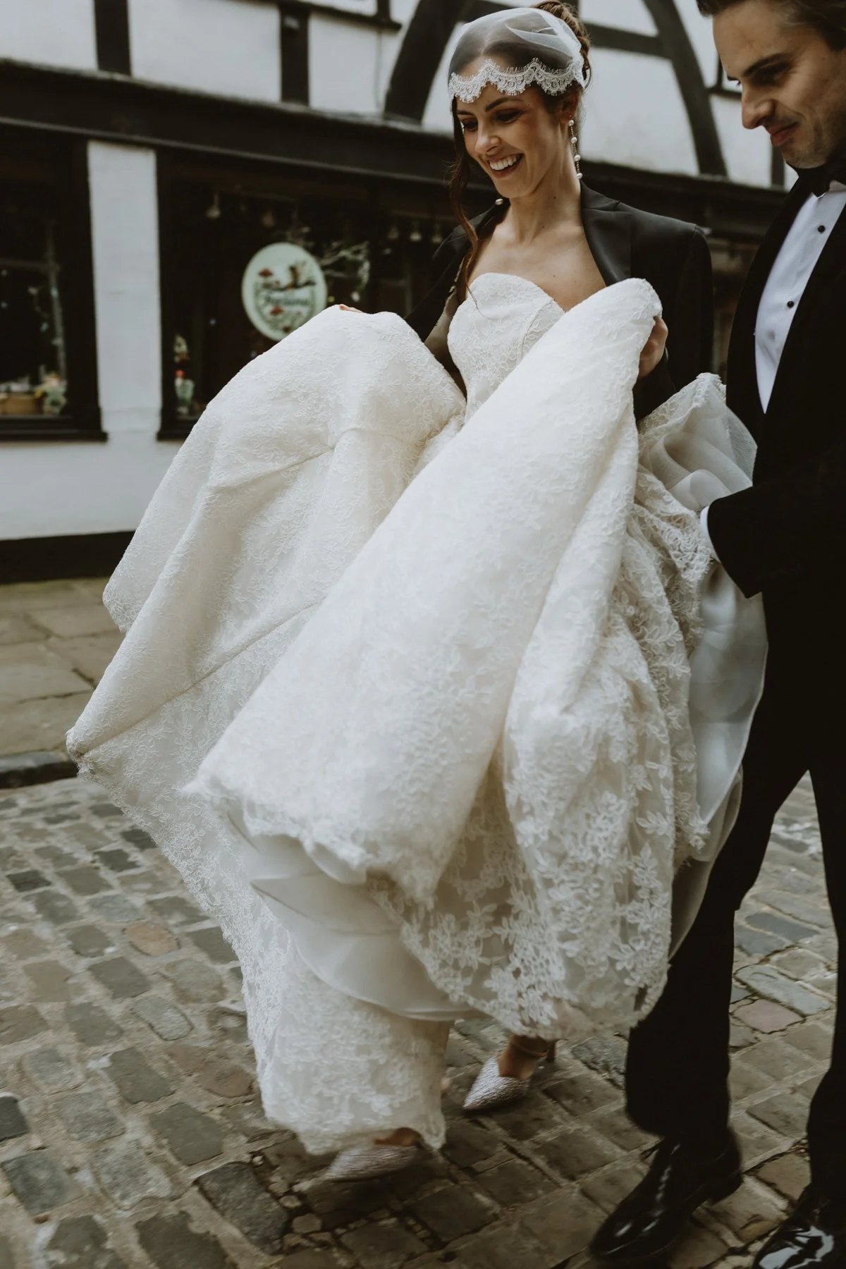 A bride in a white lace wedding dress being carried by a groom in a black tuxedo outside on a cobblestone street.