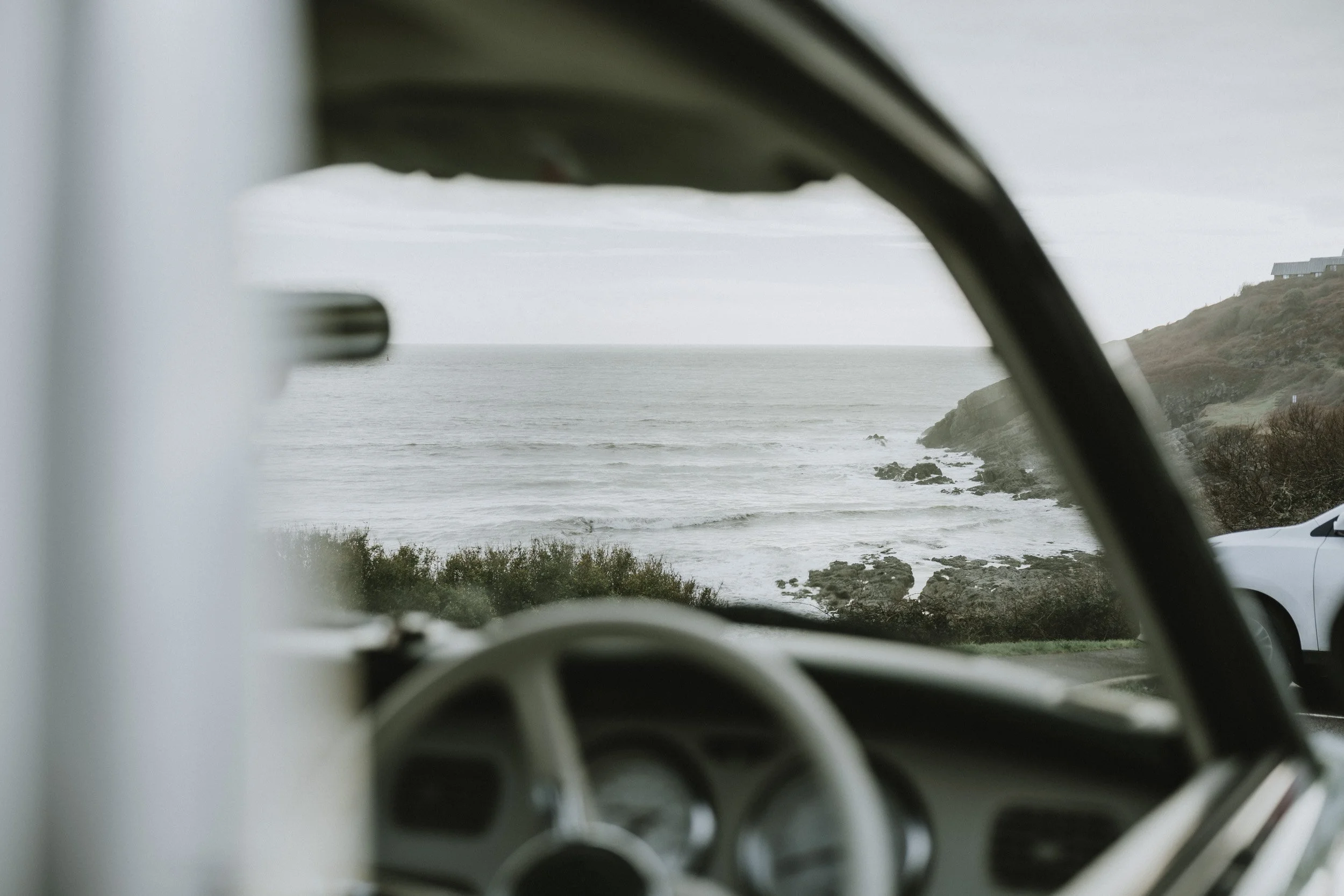 View of the ocean and coastline seen through a vehicle's side mirror, with a house on a hillside in the distance.