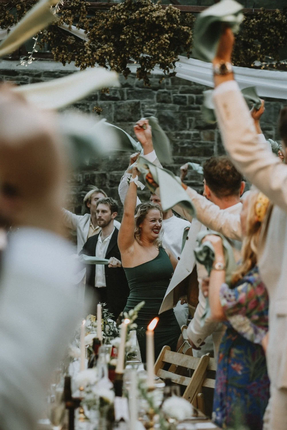 People celebrating at a wedding reception, waving napkins in the air, seated at a long decorated table with candles and flowers, with a stone wall in the background.