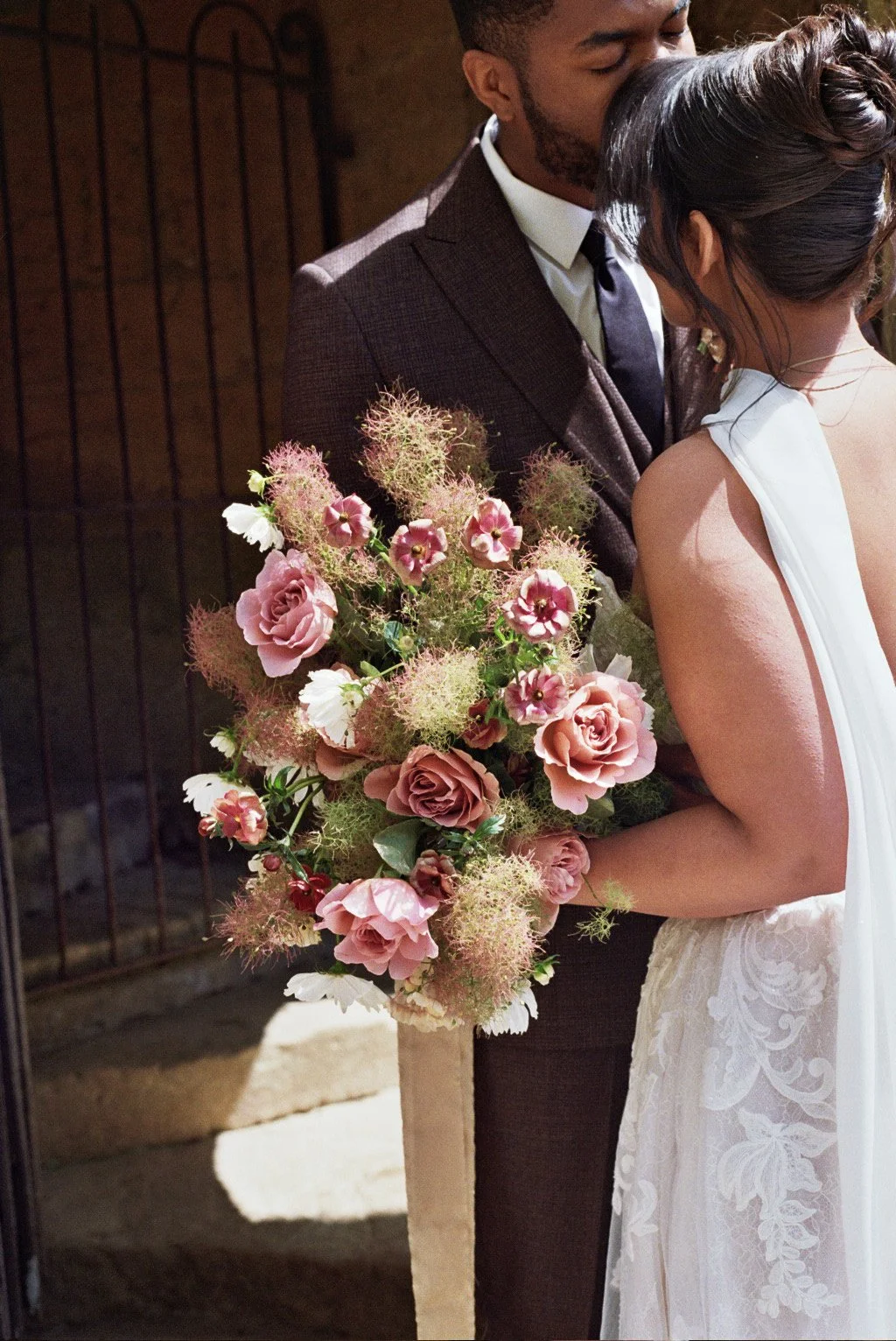 A bride and groom during their wedding ceremony, with the groom embracing the bride from behind. The bride holds a large bouquet of pink and white flowers, including roses and other blossoms. The scene is illuminated by natural light, with a dark rustic background.