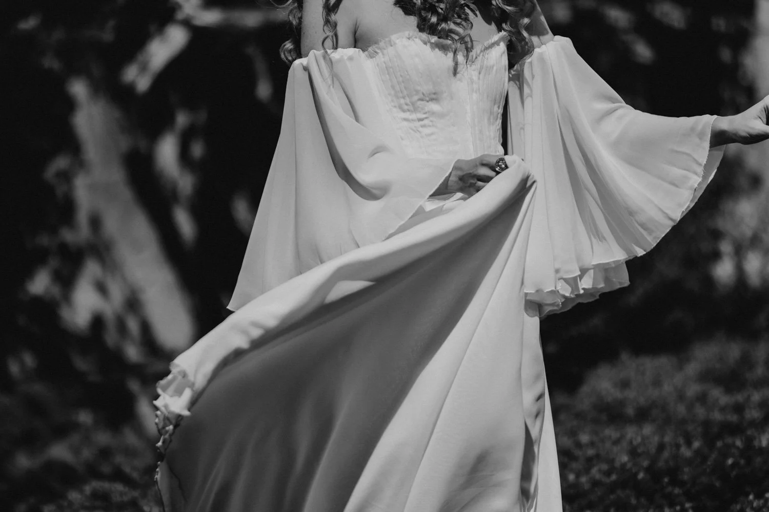 A woman in an elegant, flowing white dress holding the fabric of her gown with one hand, outdoors in a natural setting, in black and white.