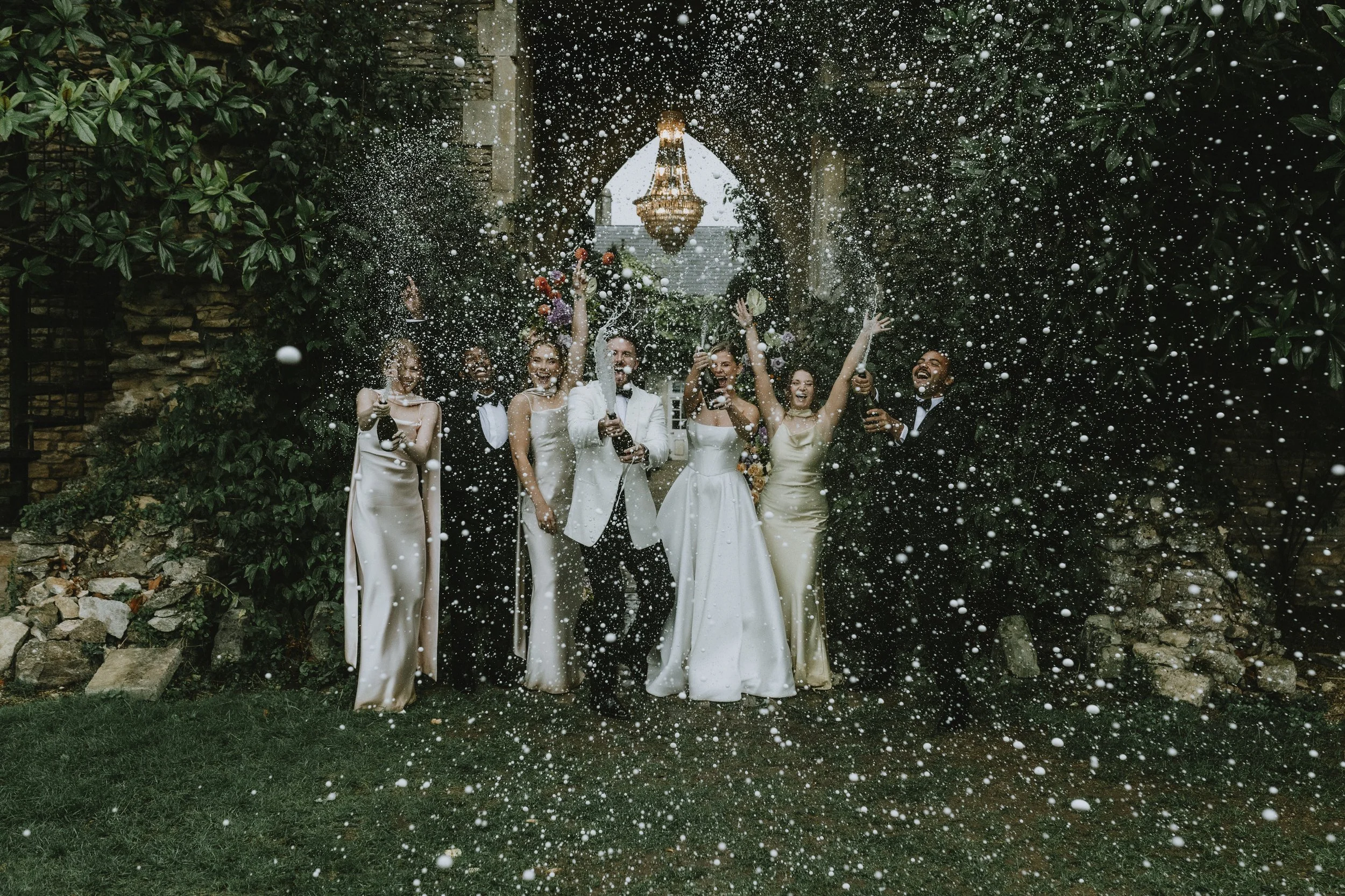 Celebrating wedding with guests spraying champagne under a garden arch, with greenery and a chandelier in the background.