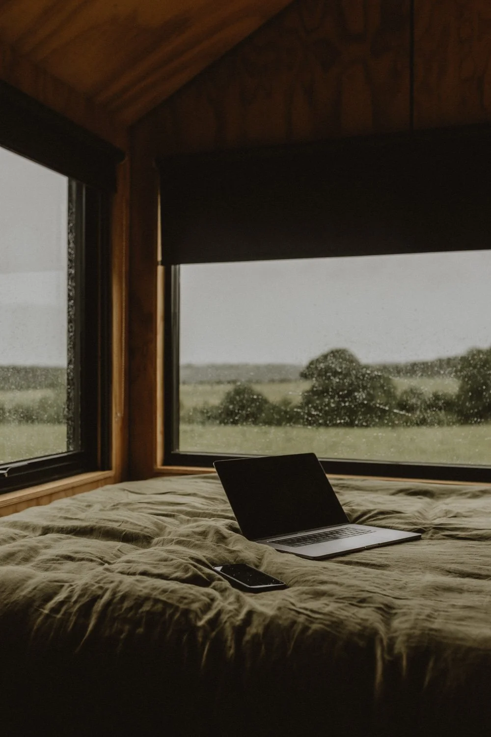 A cozy bed with a laptop and smartphone, overlooking a rainy landscape through corner windows in a wooden cabin.