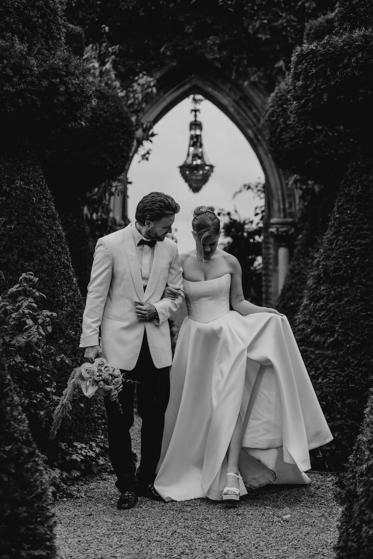A black-and-white photo of a bride and groom walking together outdoors, surrounded by lush greenery. The groom is dressed in a light-colored tuxedo with a bow tie, and the bride is wearing a strapless wedding gown with a full skirt. They are holding hands, with the bride lifting her dress slightly to reveal her shoes. An ornate arch and a hanging chandelier are visible in the background.