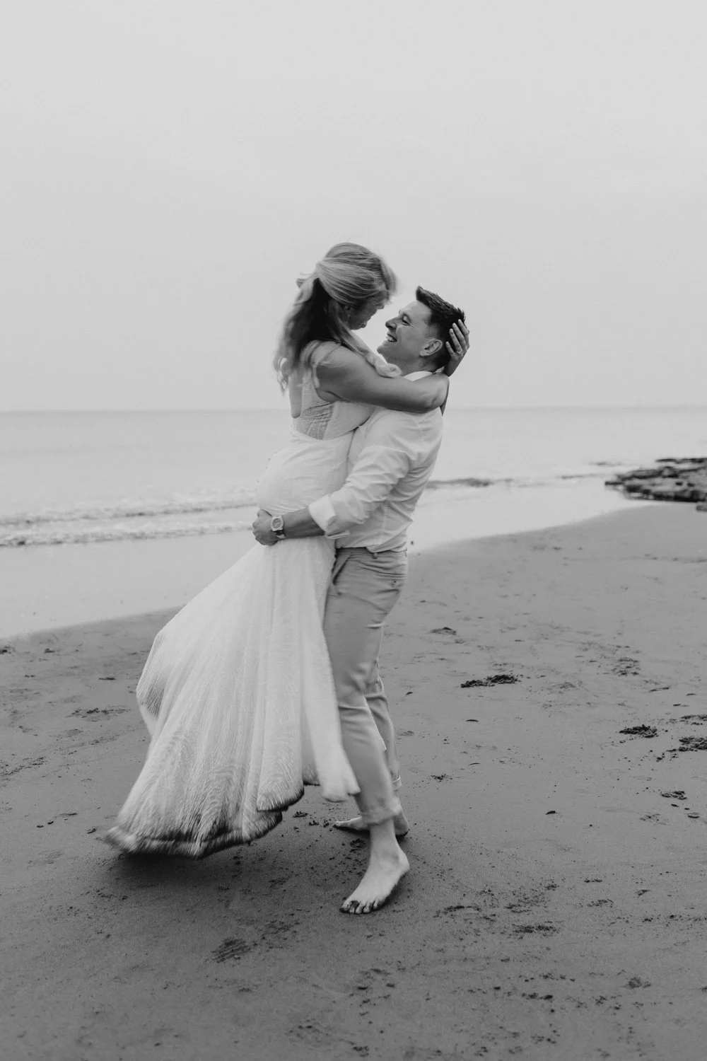 A couple on a beach, with the man lifting the woman, both smiling and embracing, in black and white.