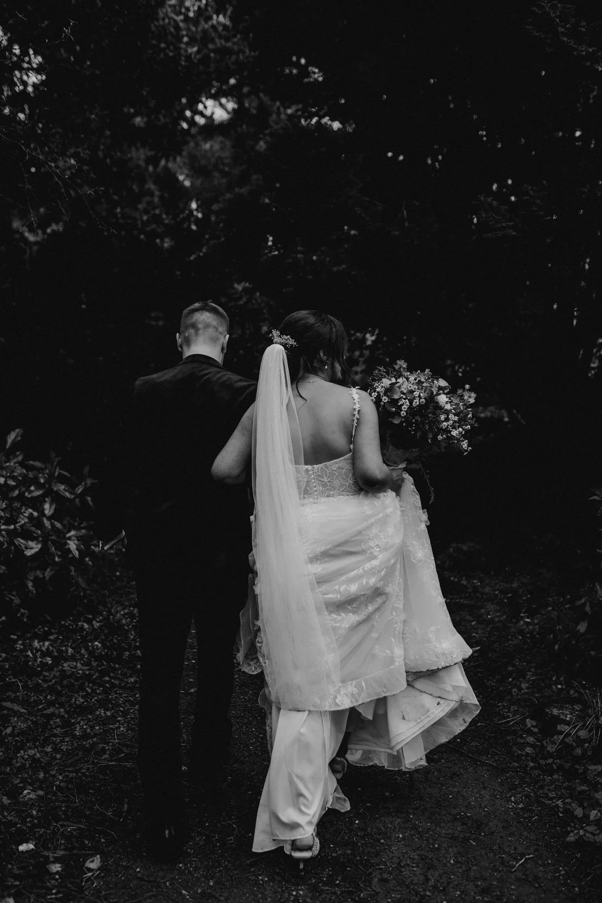 Black and white photo of a bride and groom walking away from the camera through a wooded area, with the bride holding a bouquet of flowers.
