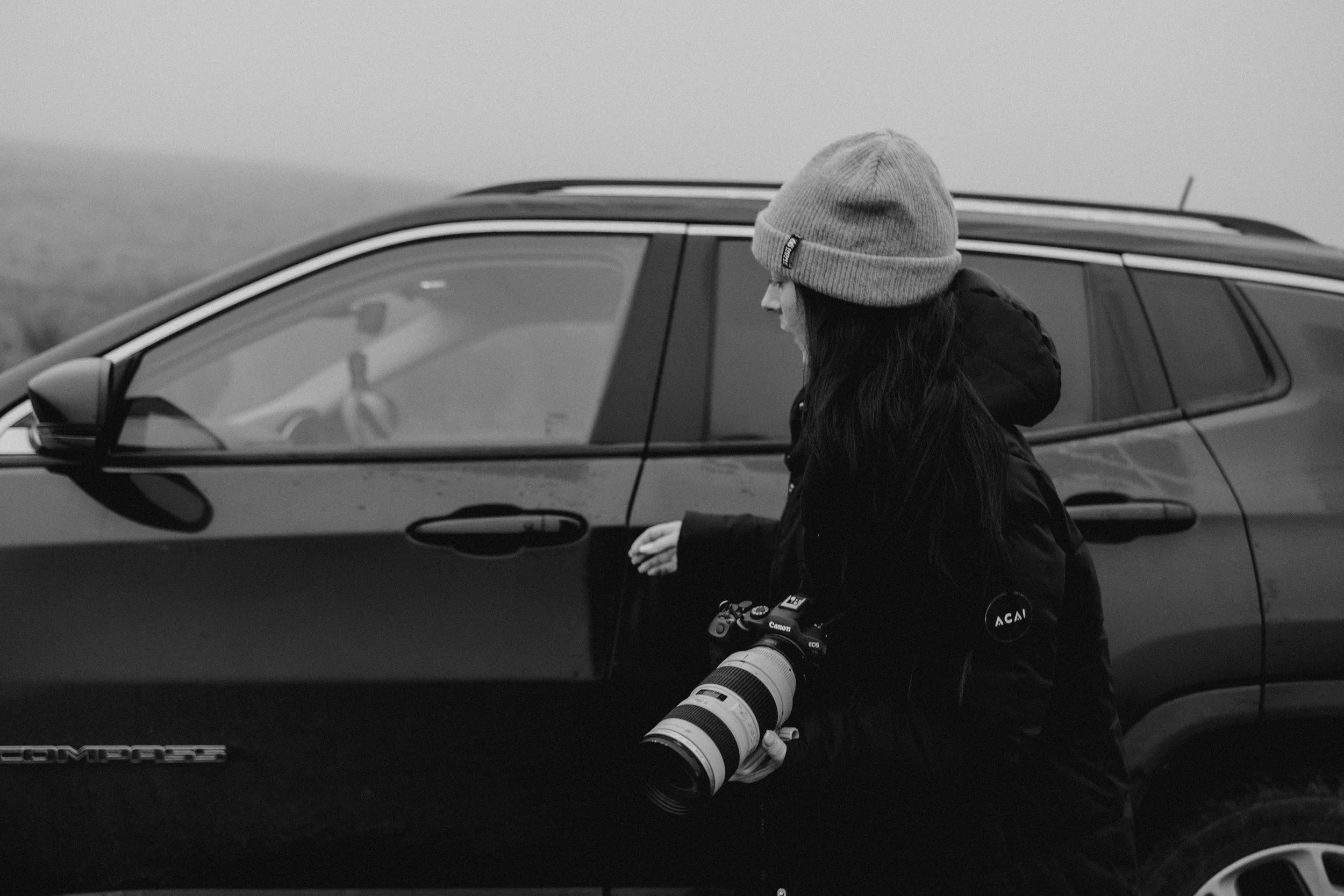 A woman with long dark hair wearing a gray beanie and black jacket standing next to a black Toyota Tacoma truck, holding a professional camera with a large telephoto lens.
