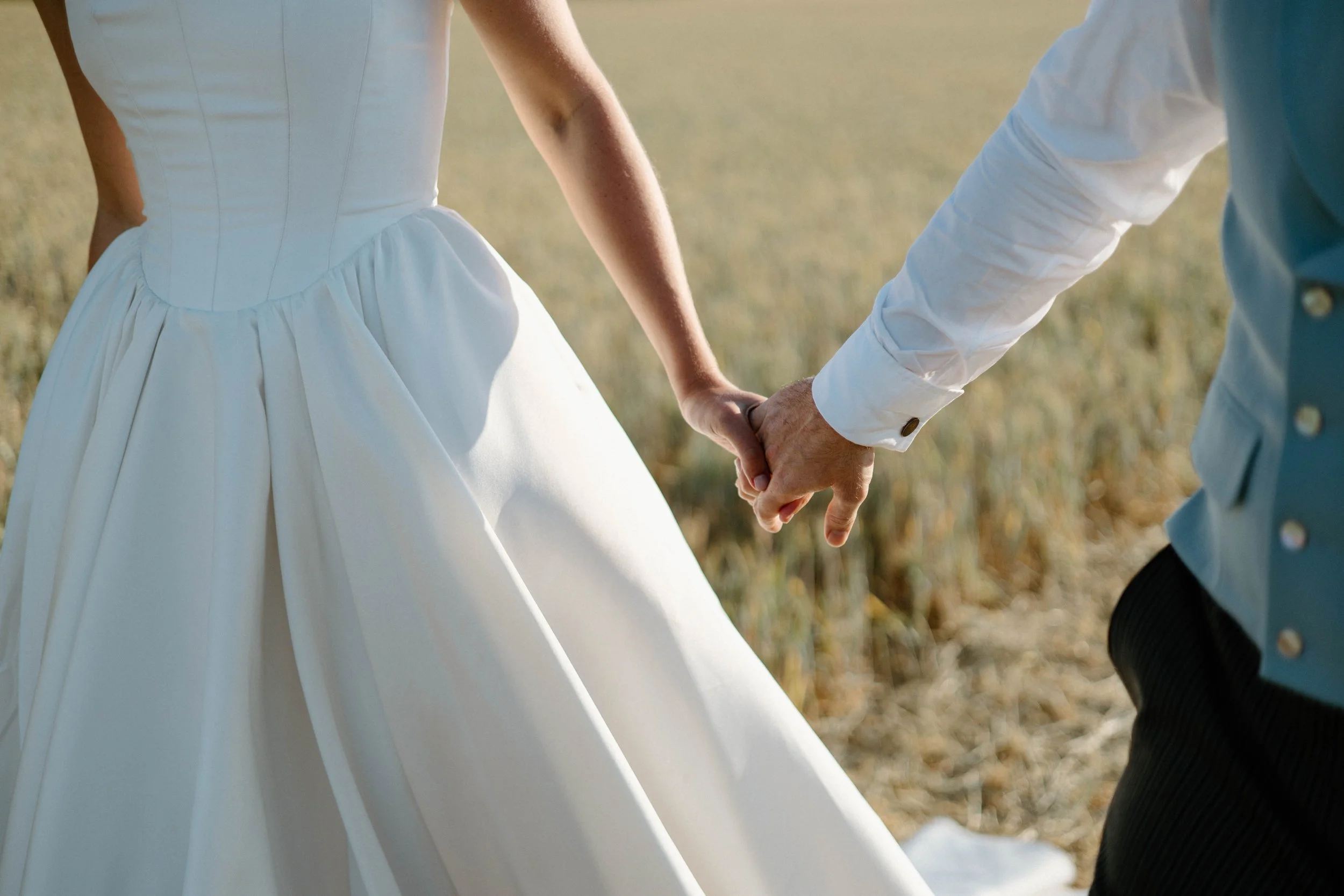 A bride and groom holding hands outdoors in a field, with the bride wearing a white wedding dress and the groom in a white shirt with a teal vest.