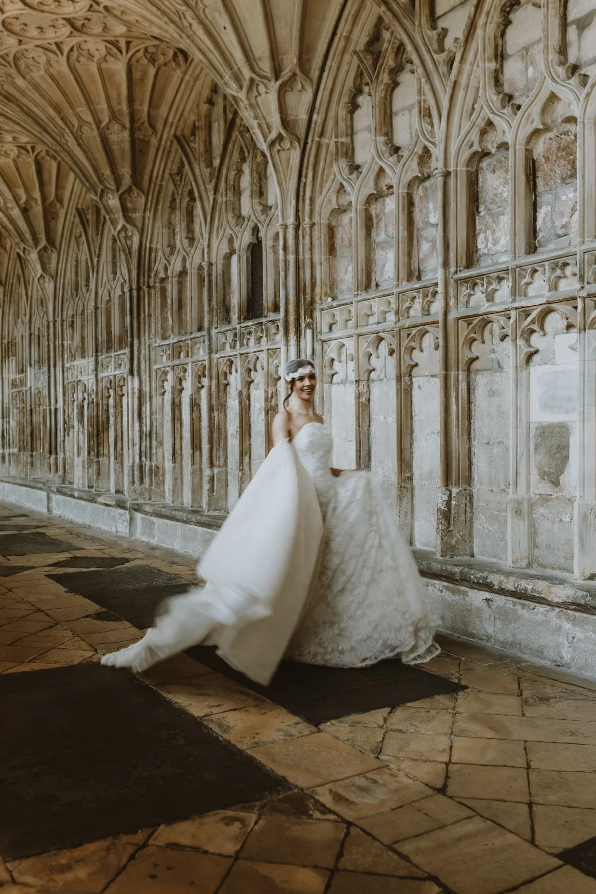 A woman in a white wedding dress walking in a historic stone corridor with vaulted gothic ceilings.