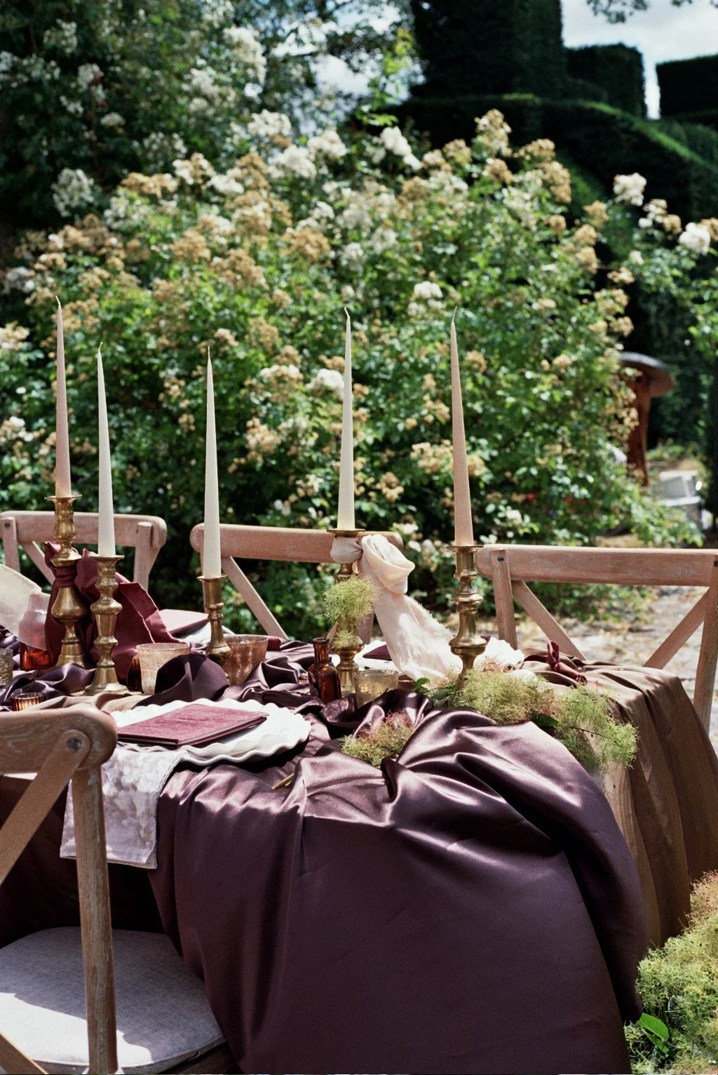 An outdoor dining table set with tall candles, cloth napkins, and decorative greenery, surrounded by wooden chairs, with a lush garden of flowering bushes and trees in the background.