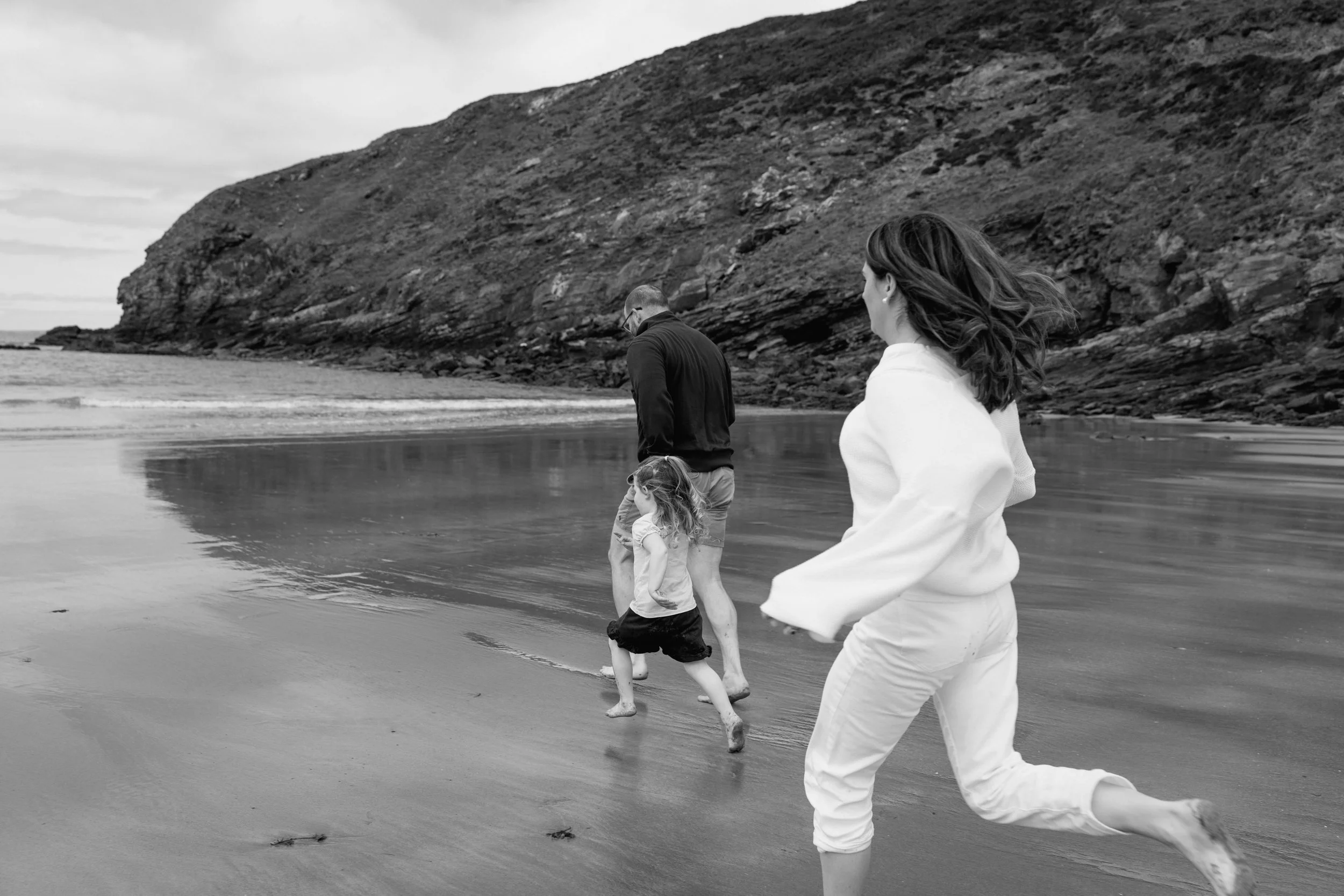 A family of three, a man, woman, and young girl, walking barefoot along the shoreline of a beach with a rocky cliff in the background, in black and white.