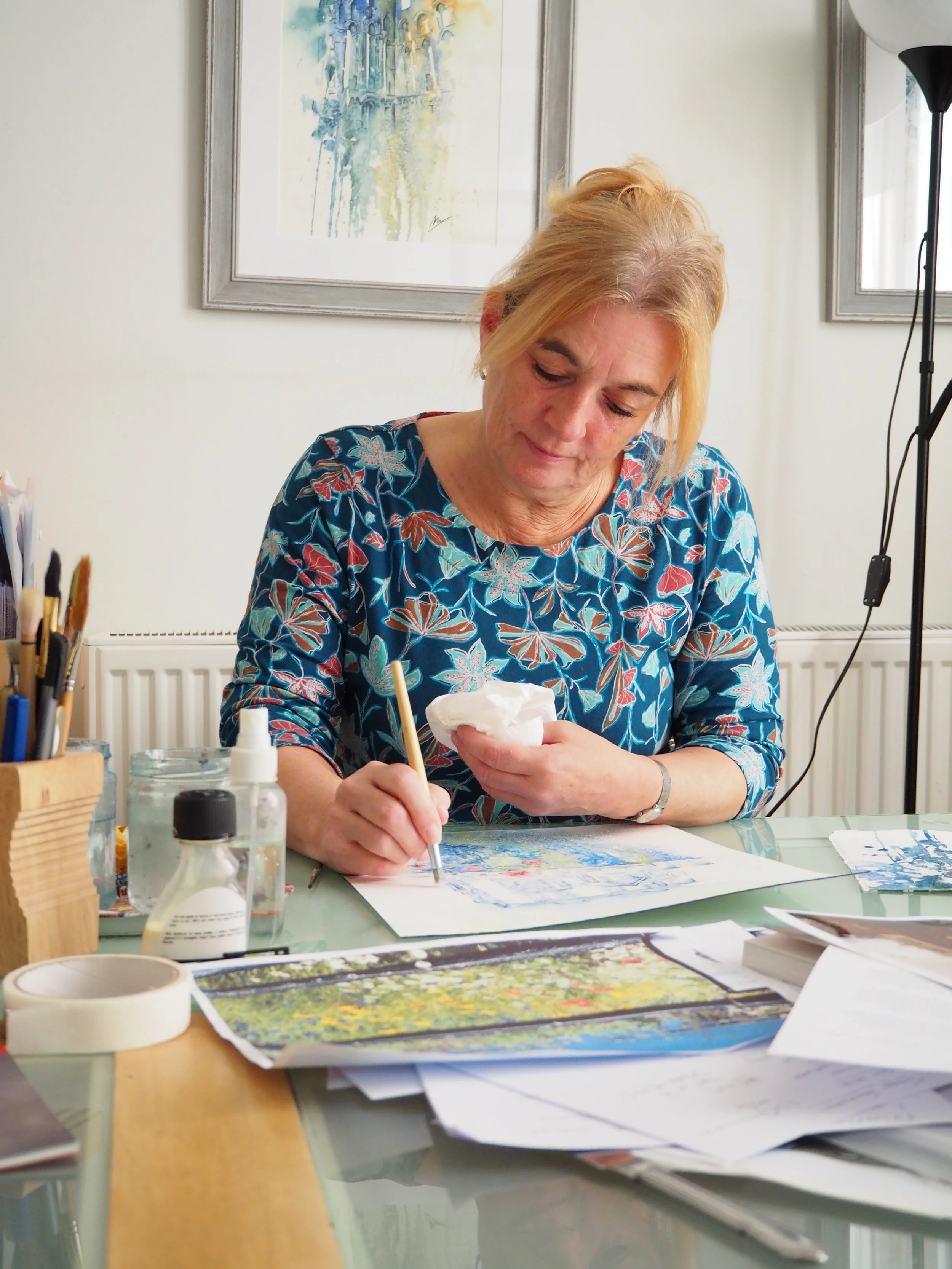 Woman painting at a workspace surrounded by brushes, watercolor supplies, and artwork
