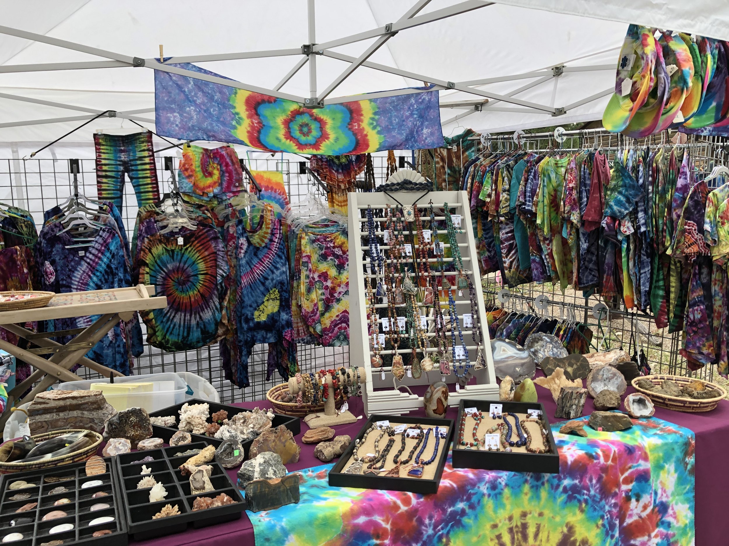 A market stall with colorful tie-dye clothing, jewelry, and mineral rocks displayed on tables and racks under a white canopy.