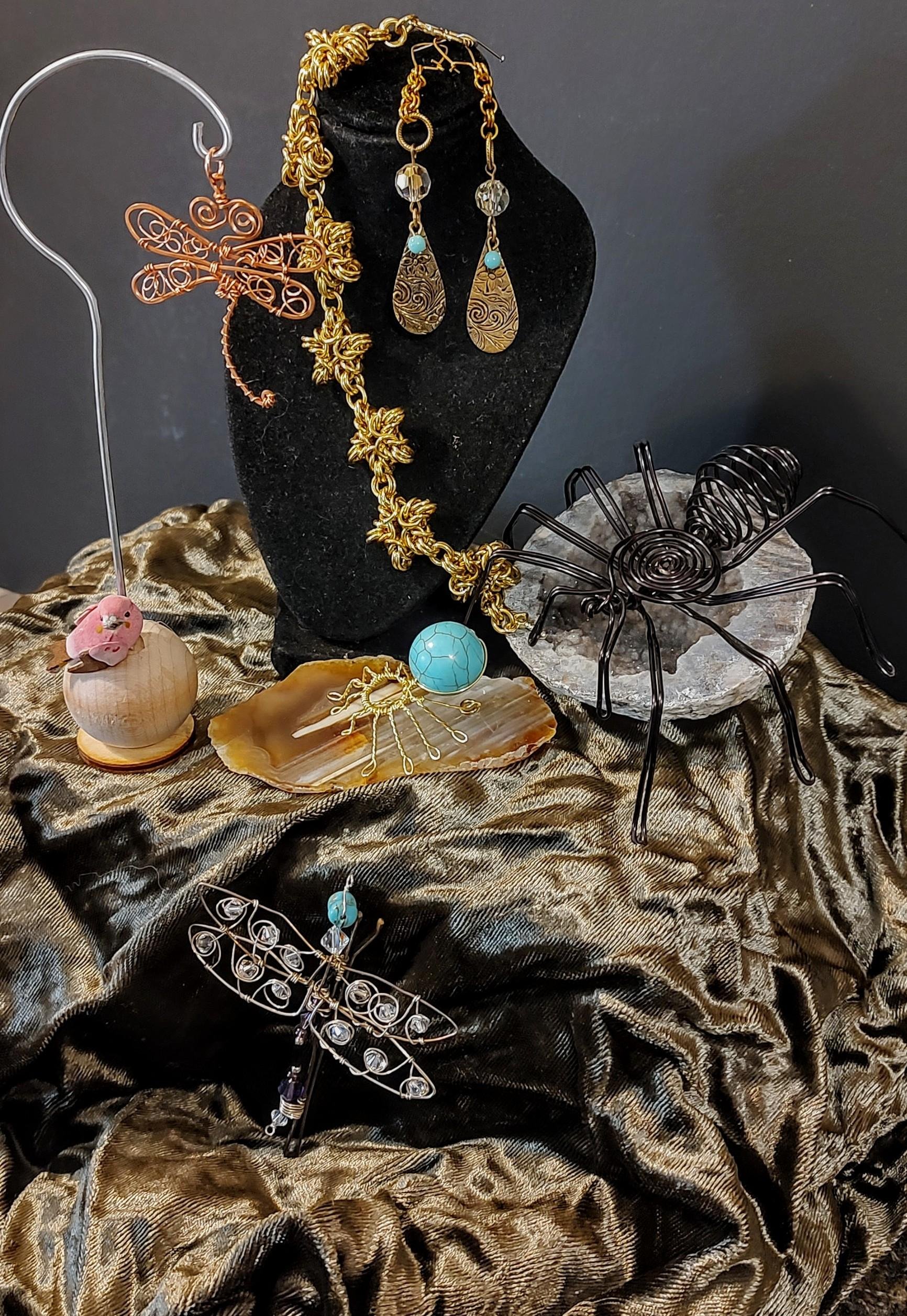 Collection of jewelry including a pair of gold earrings with teardrop-shaped engraved pendants, a gold chain necklace with floral links, a copper butterfly wire sculpture, a wire spider sculpture on a stone base, a blue patterned spherical pendant, and a gold bracelet with a radial design, all displayed on a black bust and golden fabric background.