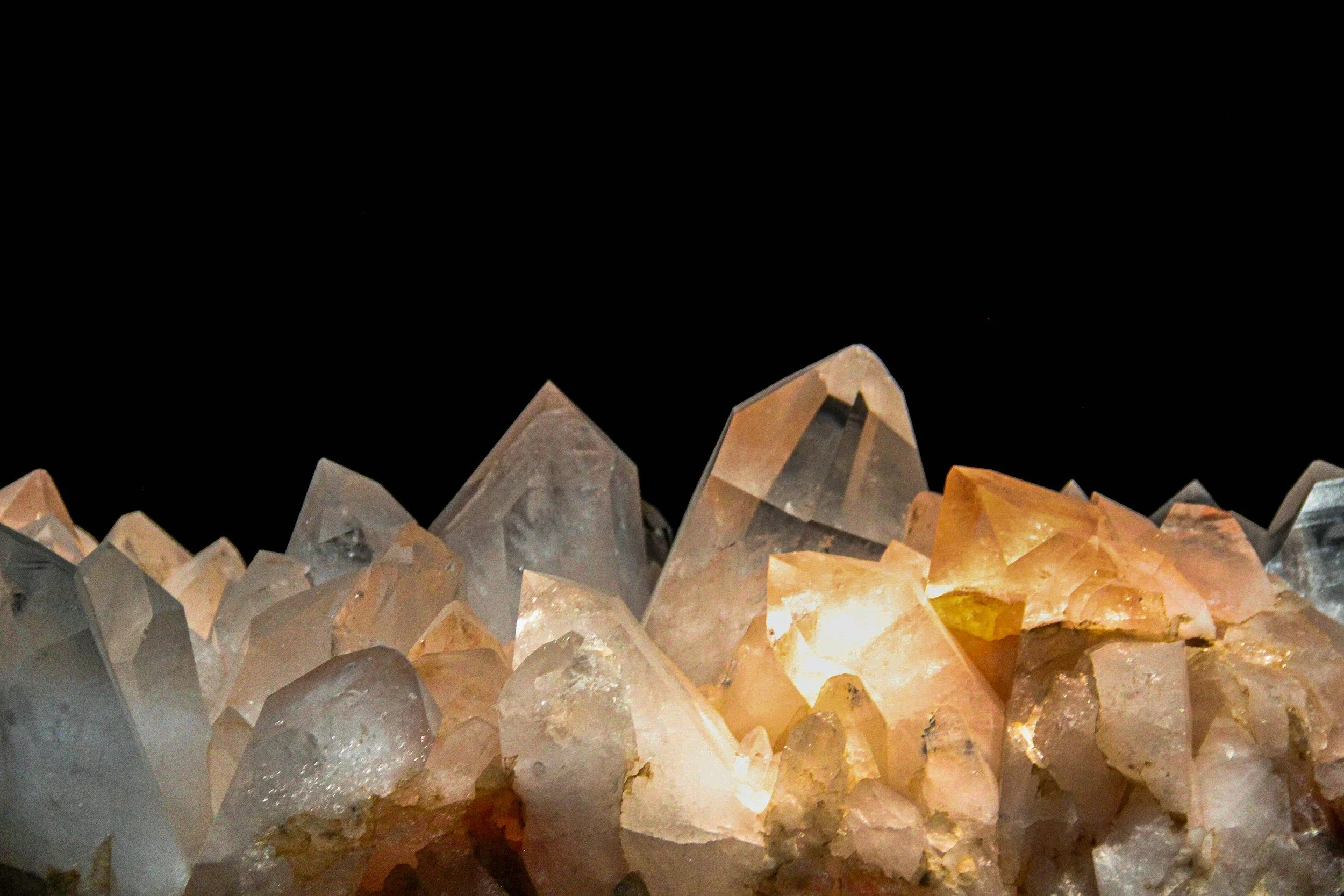 Close-up of a large cluster of illuminated quartz crystals with varying shapes and colors, against a dark background.