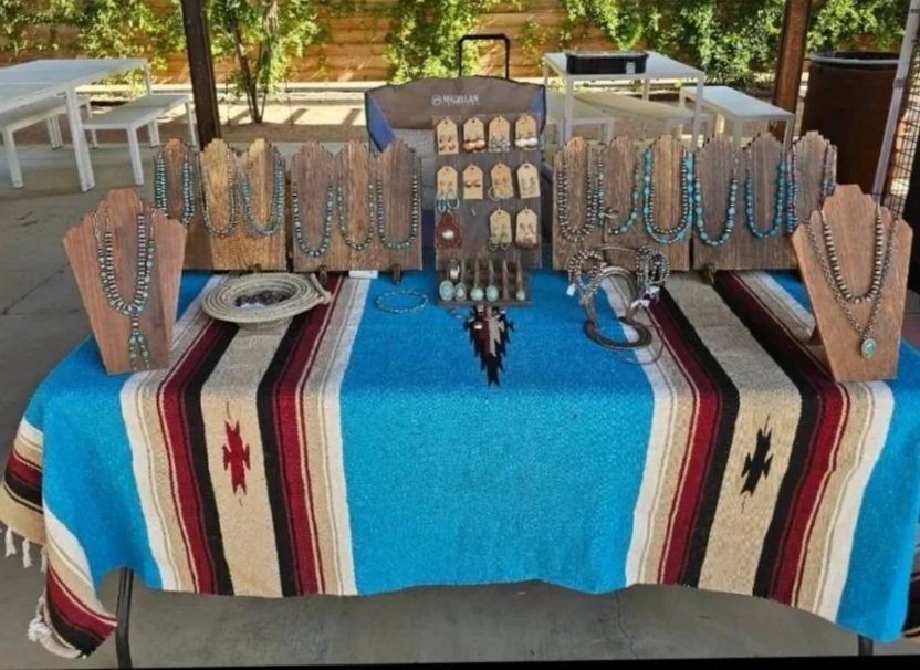 Jewelry display table with necklaces, earrings, bracelets, and pendants on wooden stands and a fabric tablecloth with southwestern patterns, set outdoors on a patio.
