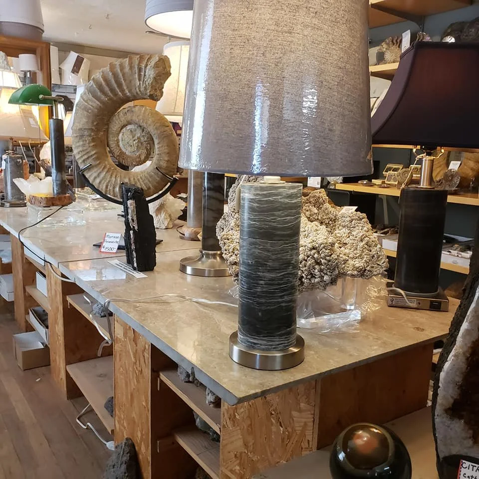 A display table in an antique or home decor shop featuring various lamps, decorative stones, and fossils, with shelves in the background holding additional items.