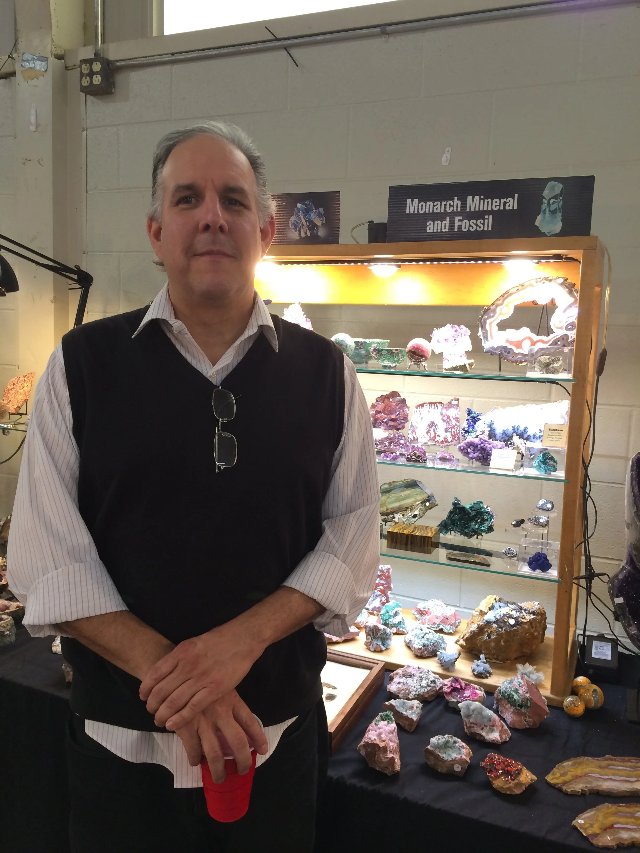 A man standing in front of a display case with various mineral and fossil specimens, including colorful rocks and crystals. A sign above the display reads 'Monarch Mineral and Fossil'.