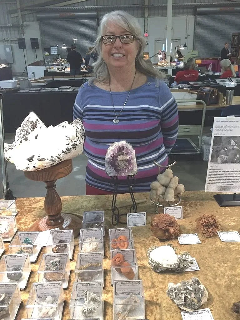 A woman with glasses and gray hair standing behind a display of various mineral and crystal specimens at an indoor exhibition or market.
