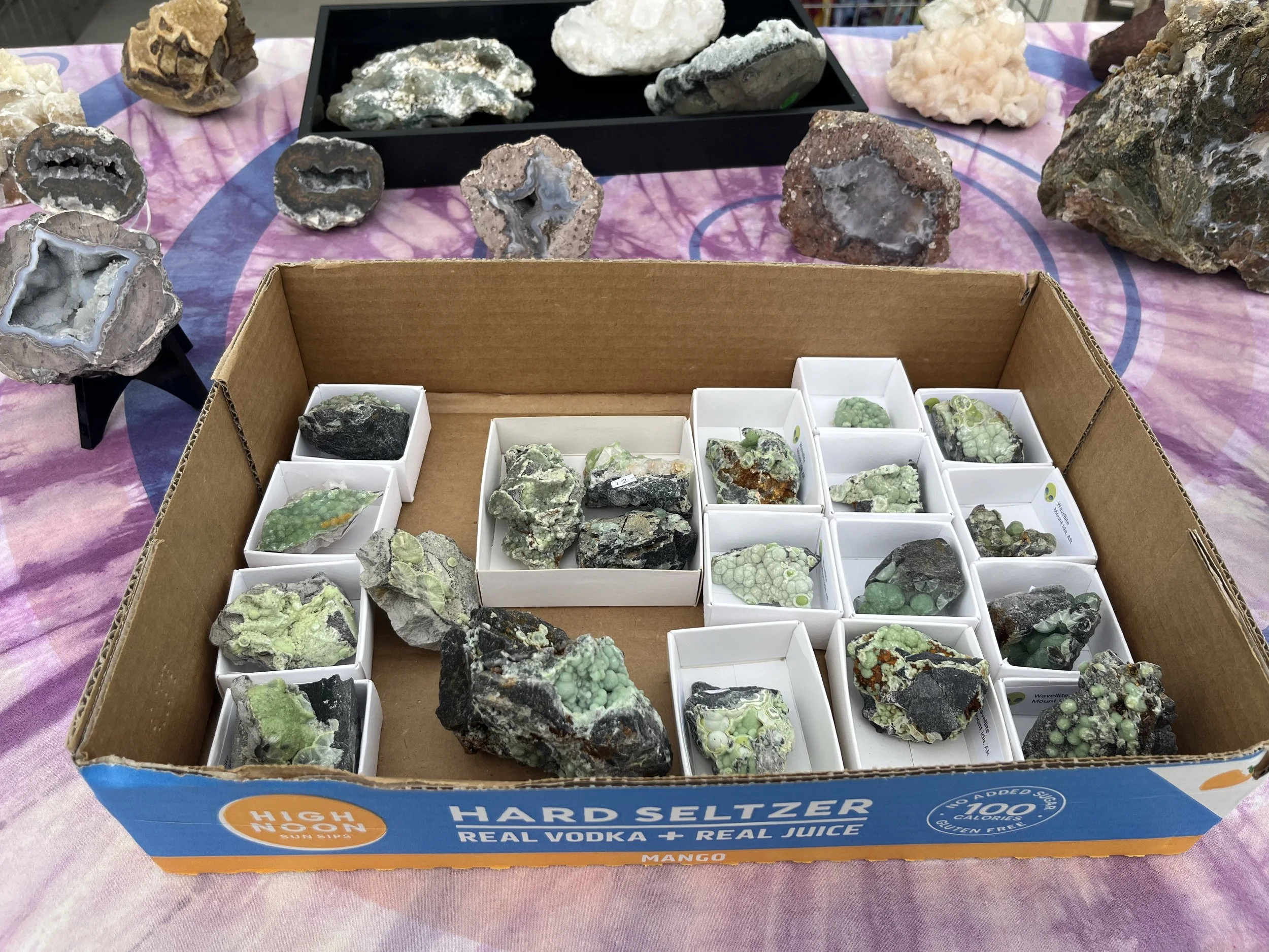Various mineral specimens displayed on a table, with a cardboard box in the foreground containing smaller rocks or minerals in white containers, some labeled with green and black text.