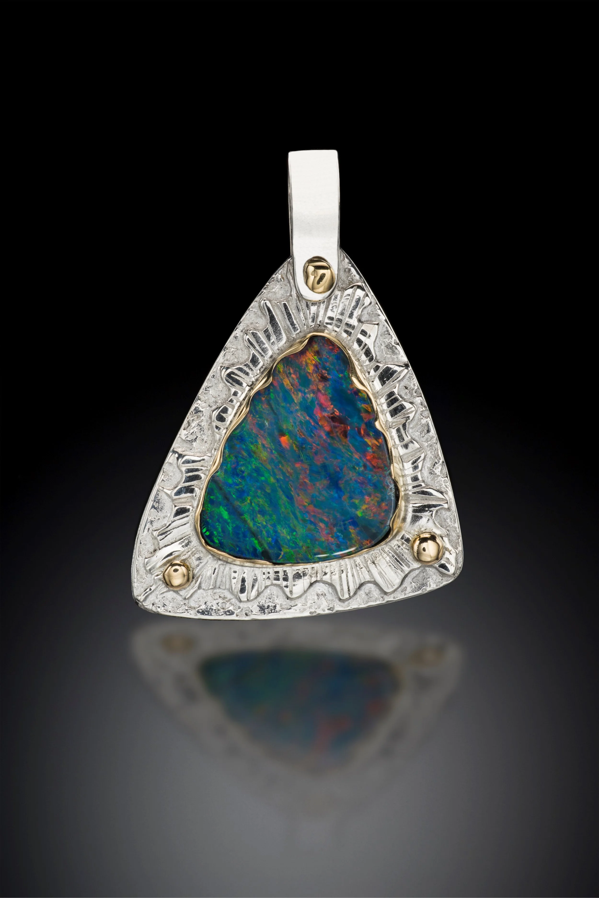 A triangular silver pendant featuring an iridescent opal stone and decorative gold accents, set against a black background with a reflection below.