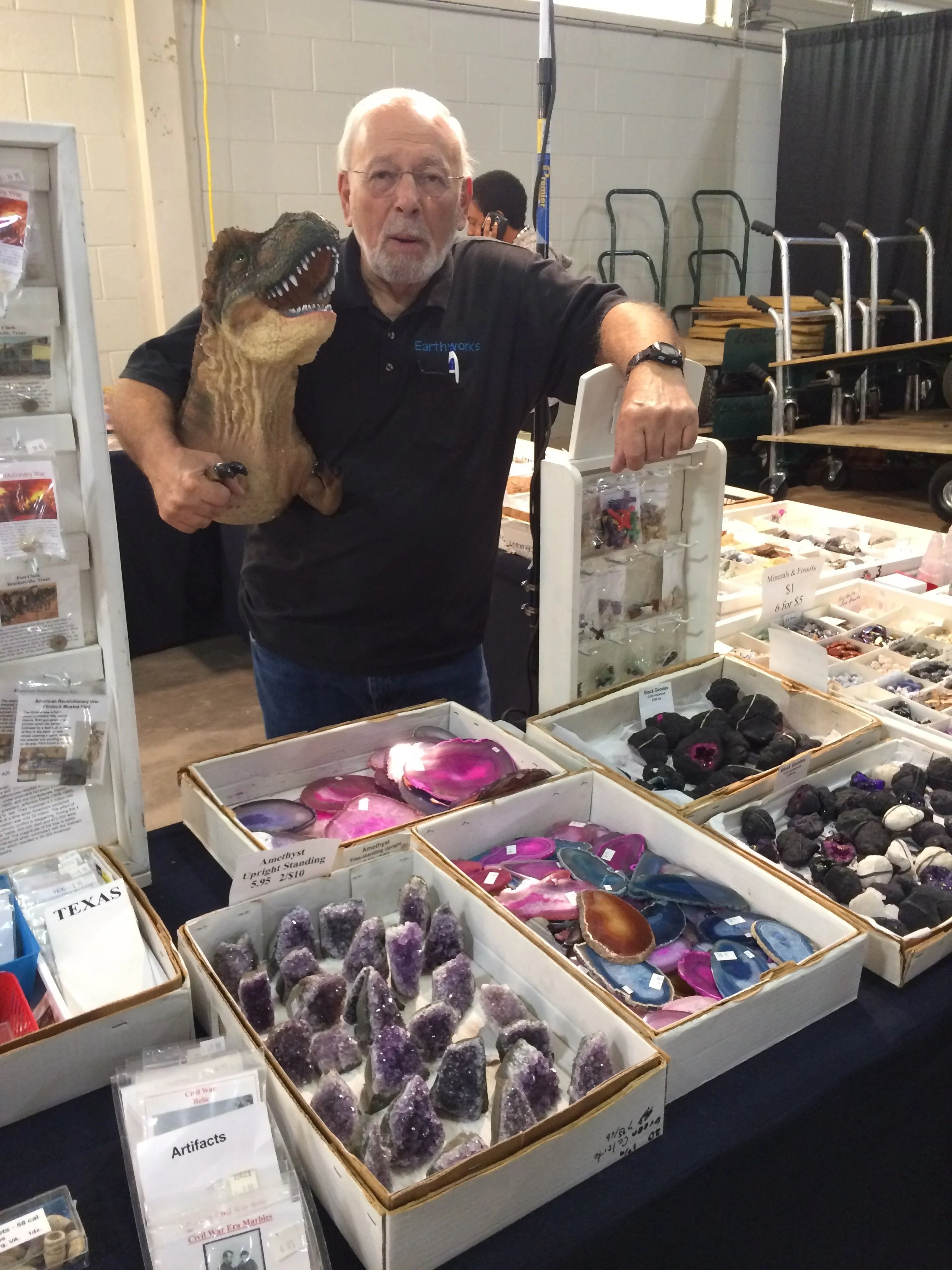 A man holding a model of a dinosaur at a table with various rocks and minerals, with shelves and carts in the background.
