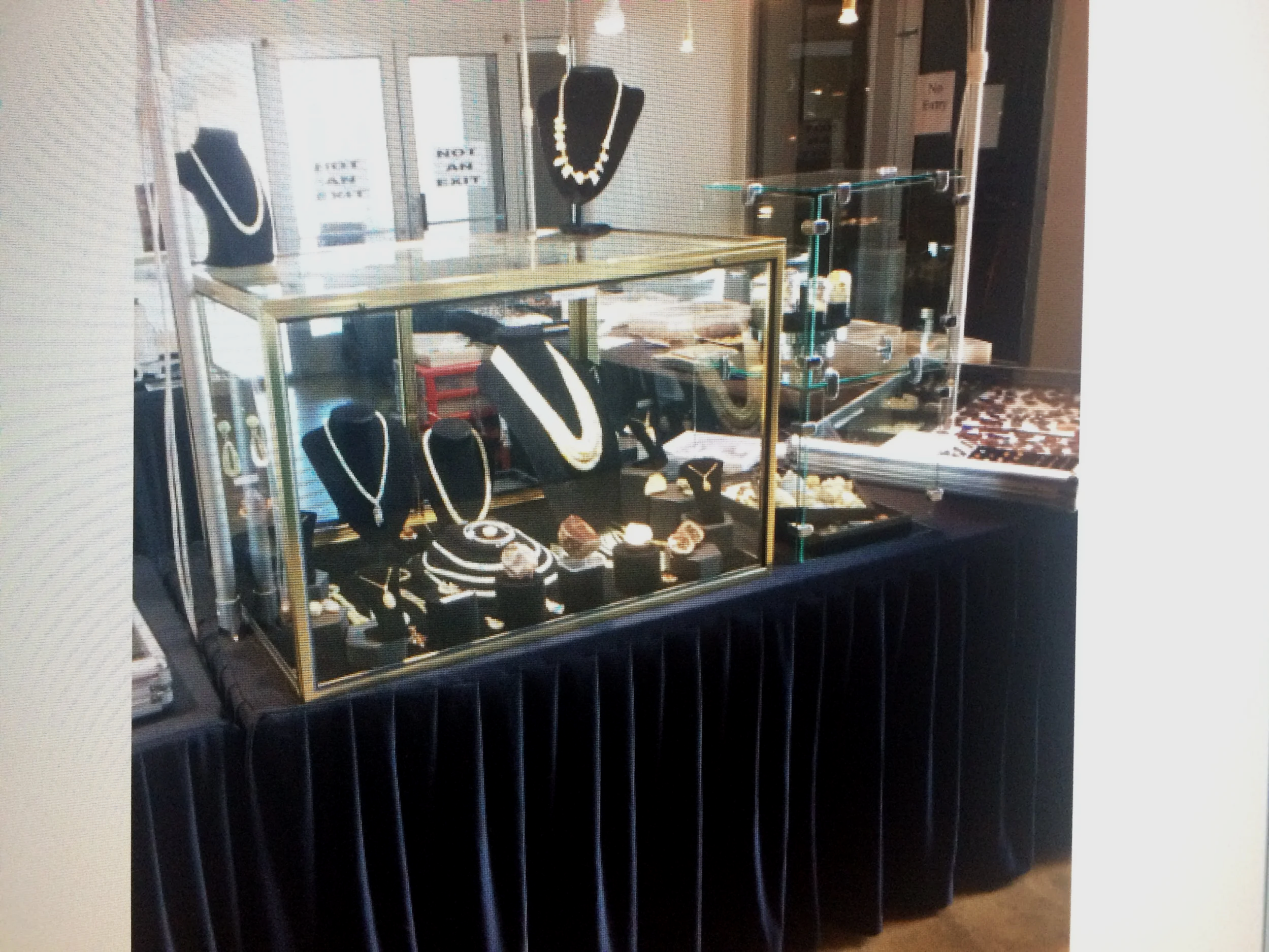 Jewelry displayed in a glass showcase, including necklaces and rings, inside a store with signs on the door that say "HOT AND DONE".