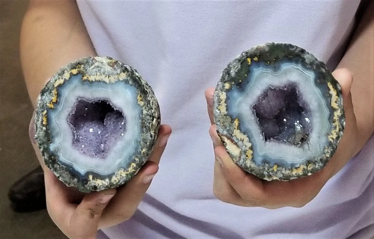 Person holding two slices of polished geodes, showing purple crystal interiors