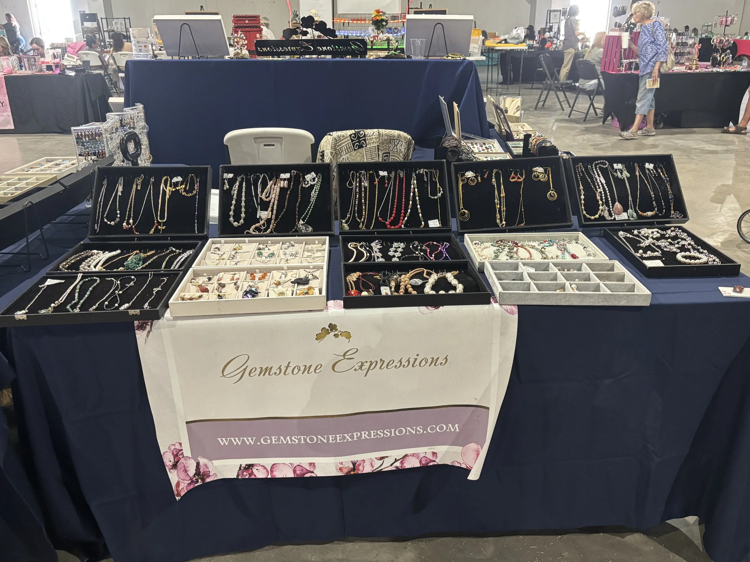 Jewelry display table with necklaces, bracelets, and rings at Gemstone Expressions vendor booth in an indoor market.