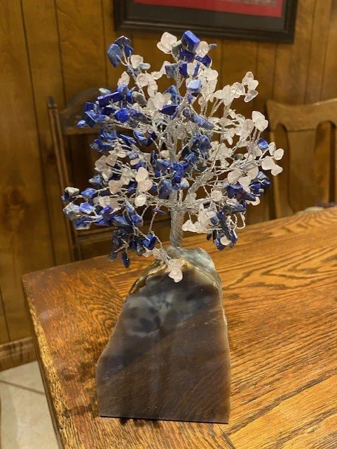 A decorative wire tree with artificial blue and white leaves, mounted on a polished stone base, placed on a wooden table.