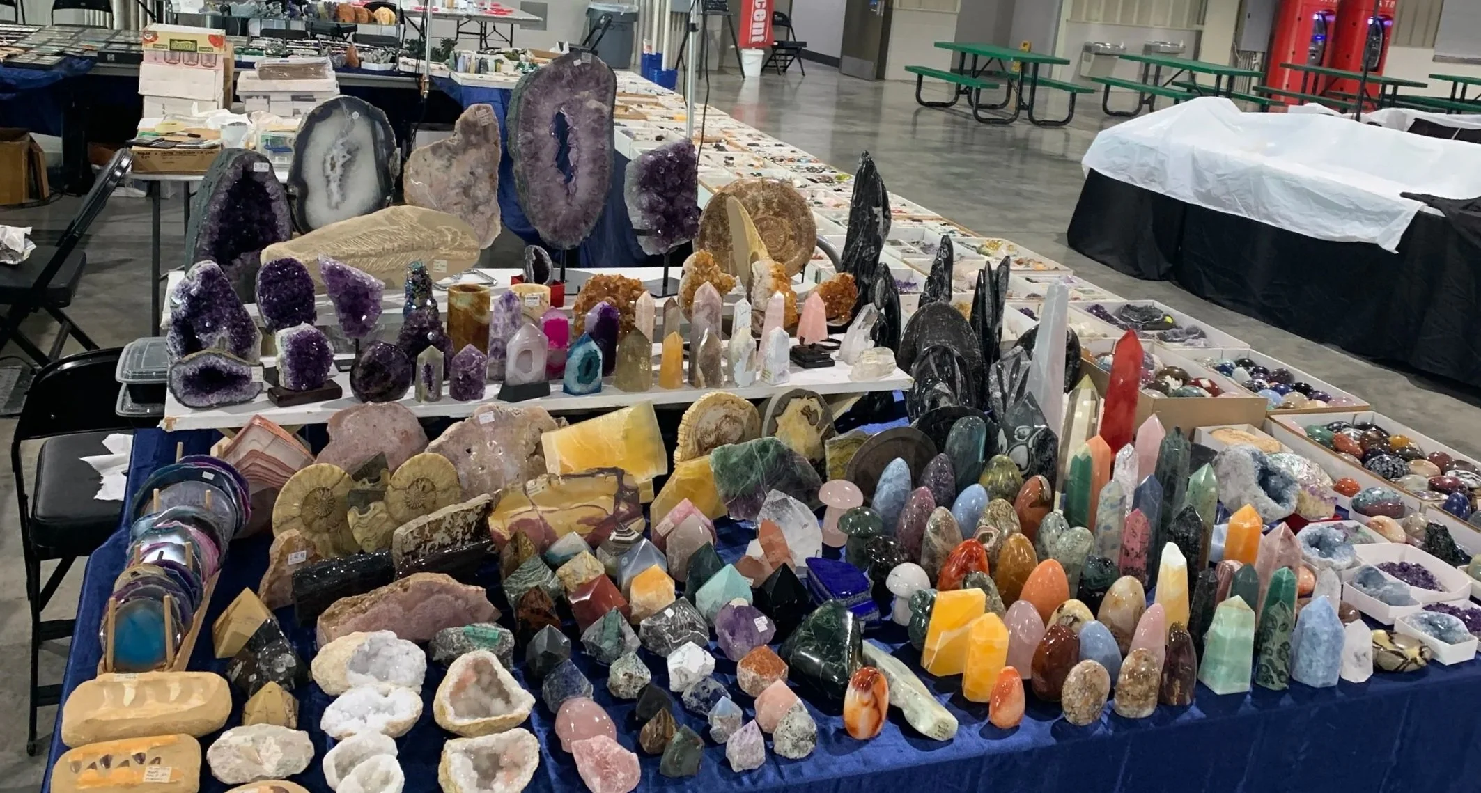 Various geodes, crystals, and polished stones displayed on tables at a market or fair, with additional stones and mineral specimens visible in the background.