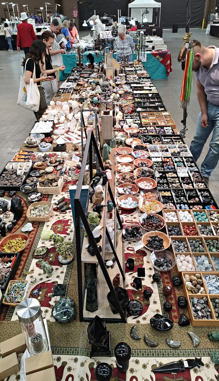 View of a large indoor flea market or craft fair with booths displaying various decorative and collectible items, including stones, minerals, figurines, jewelry, and ornaments.